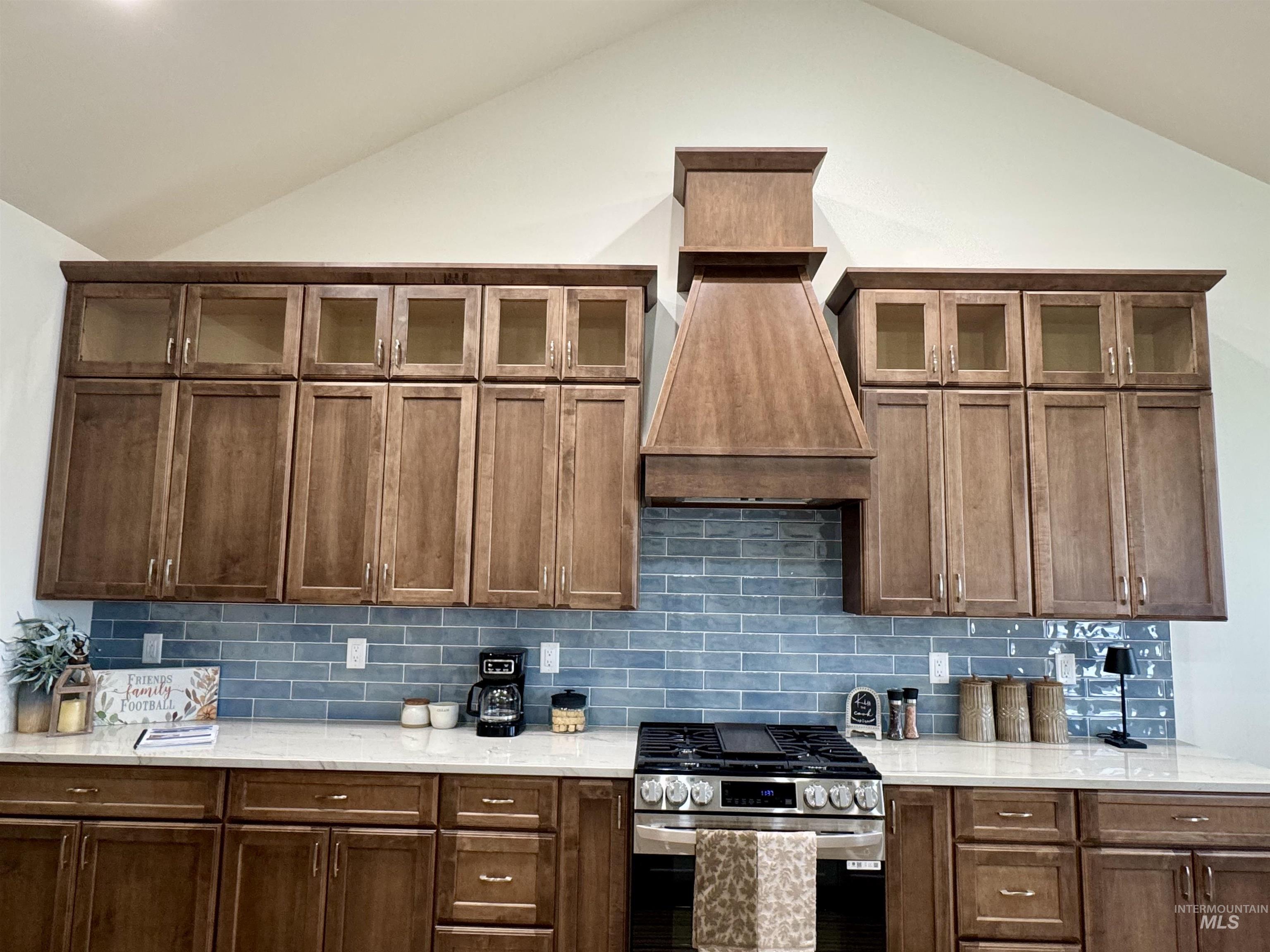 Kitchen with glass insert cabinets, stainless steel gas stove, decorative backsplash, lofted ceiling, and light stone counters