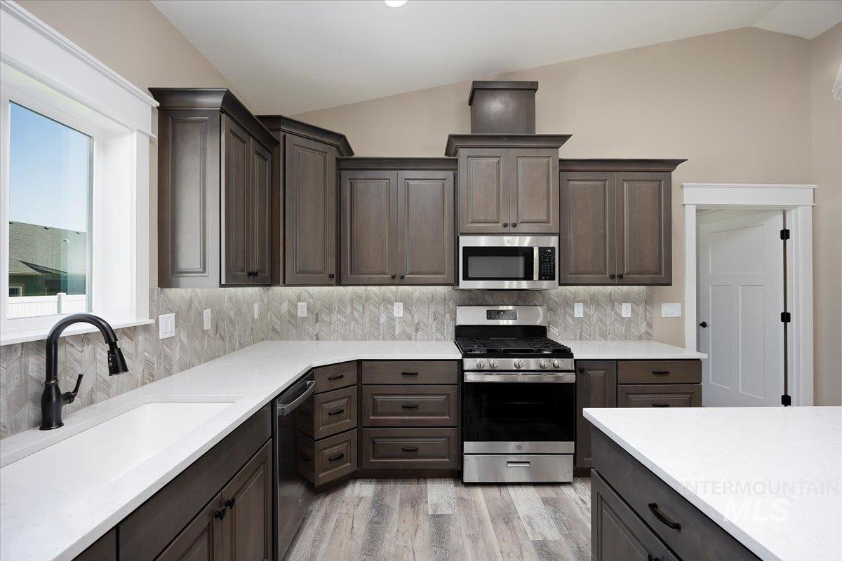 Kitchen featuring appliances with stainless steel finishes, vaulted ceiling, decorative backsplash, dark brown cabinets, and light wood finished floors