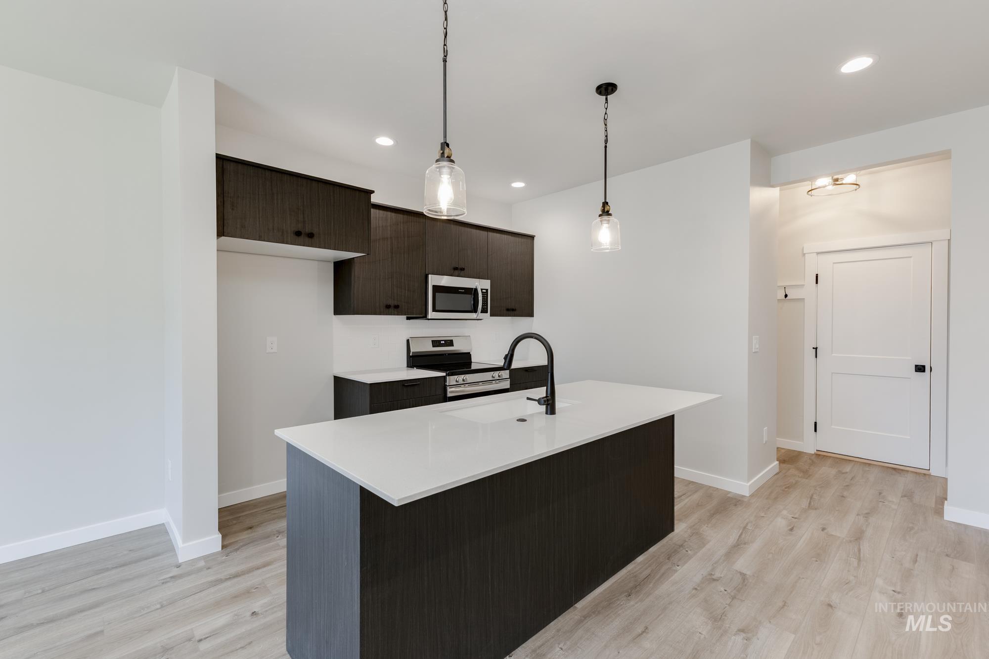 Kitchen featuring light wood-style flooring, appliances with stainless steel finishes, light countertops, a kitchen island with sink, and pendant lighting