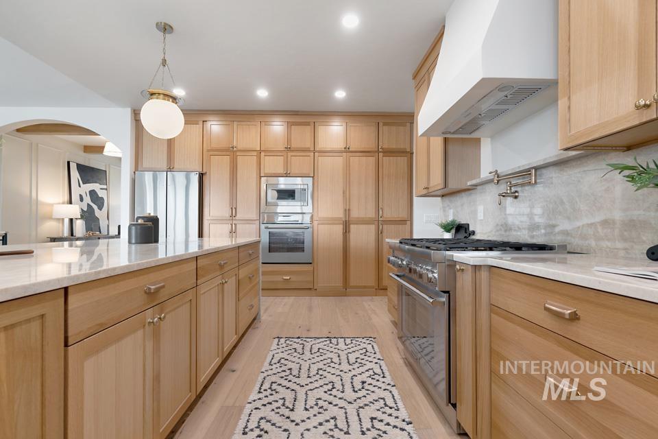 Kitchen featuring wall chimney range hood, light stone countertops, arched walkways, appliances with stainless steel finishes, and light brown cabinetry