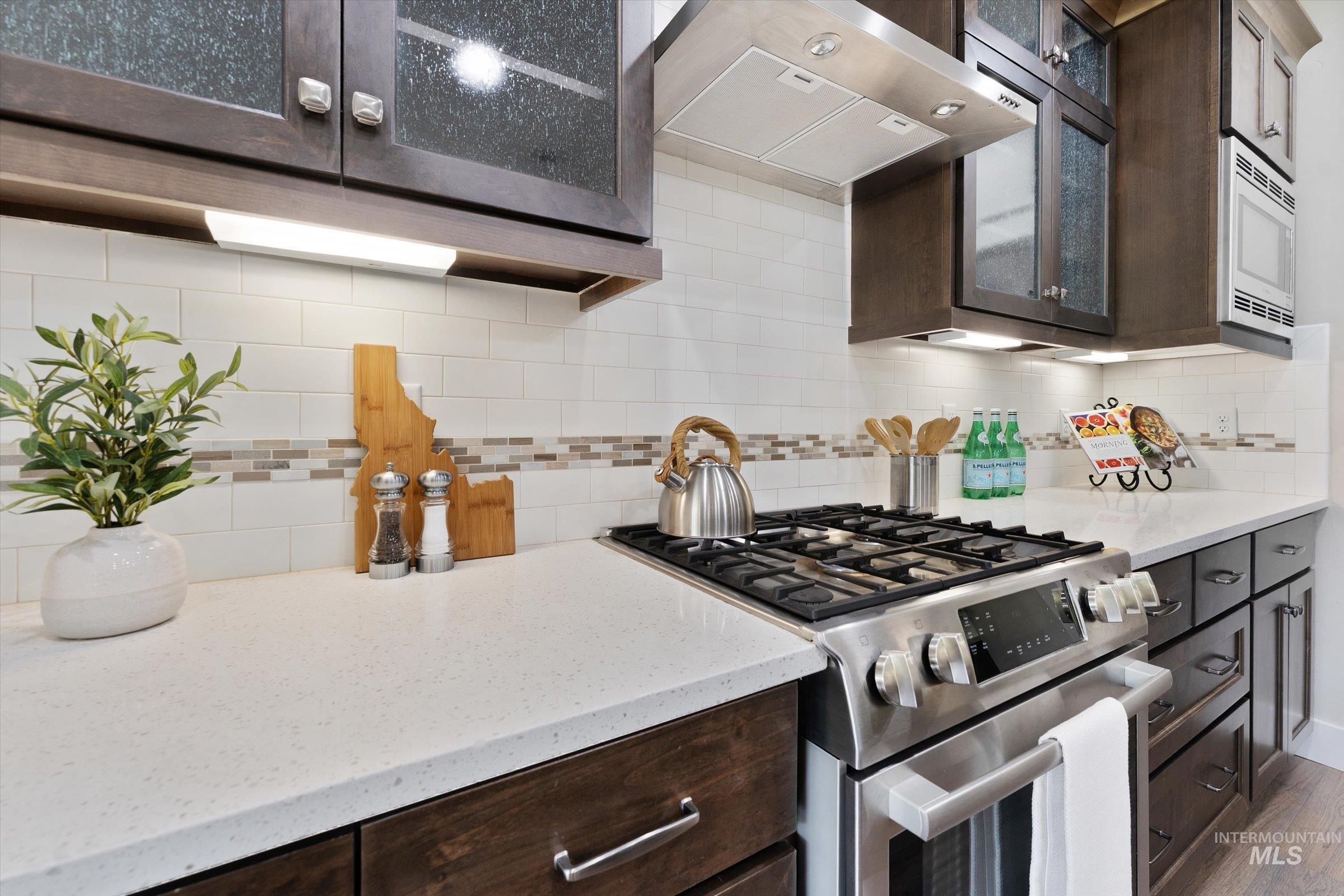 Kitchen with appliances with stainless steel finishes, glass insert cabinets, ventilation hood, light stone counters, and dark brown cabinets