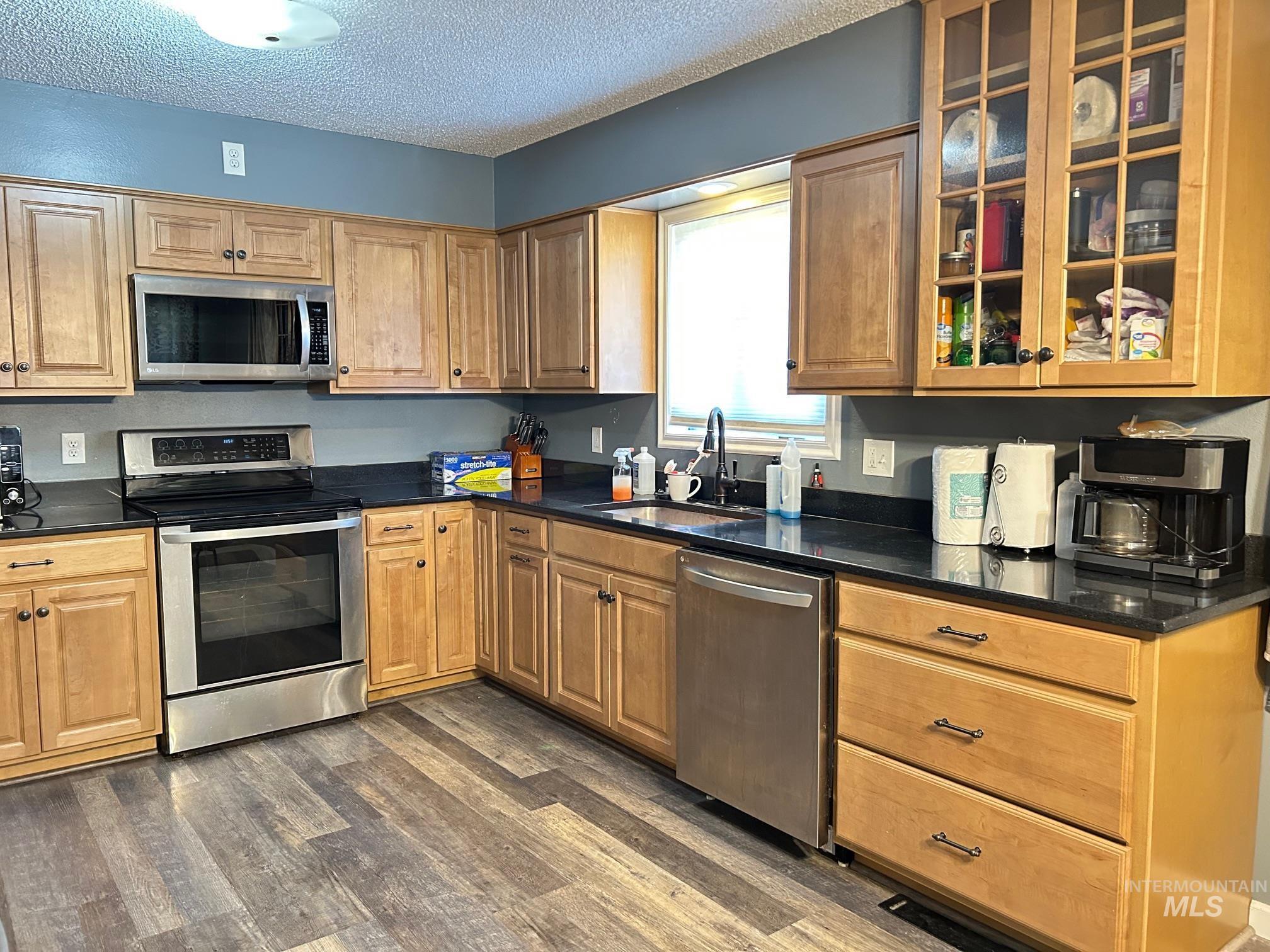 Kitchen featuring appliances with stainless steel finishes, a textured ceiling, dark countertops, dark wood finished floors, and glass insert cabinets