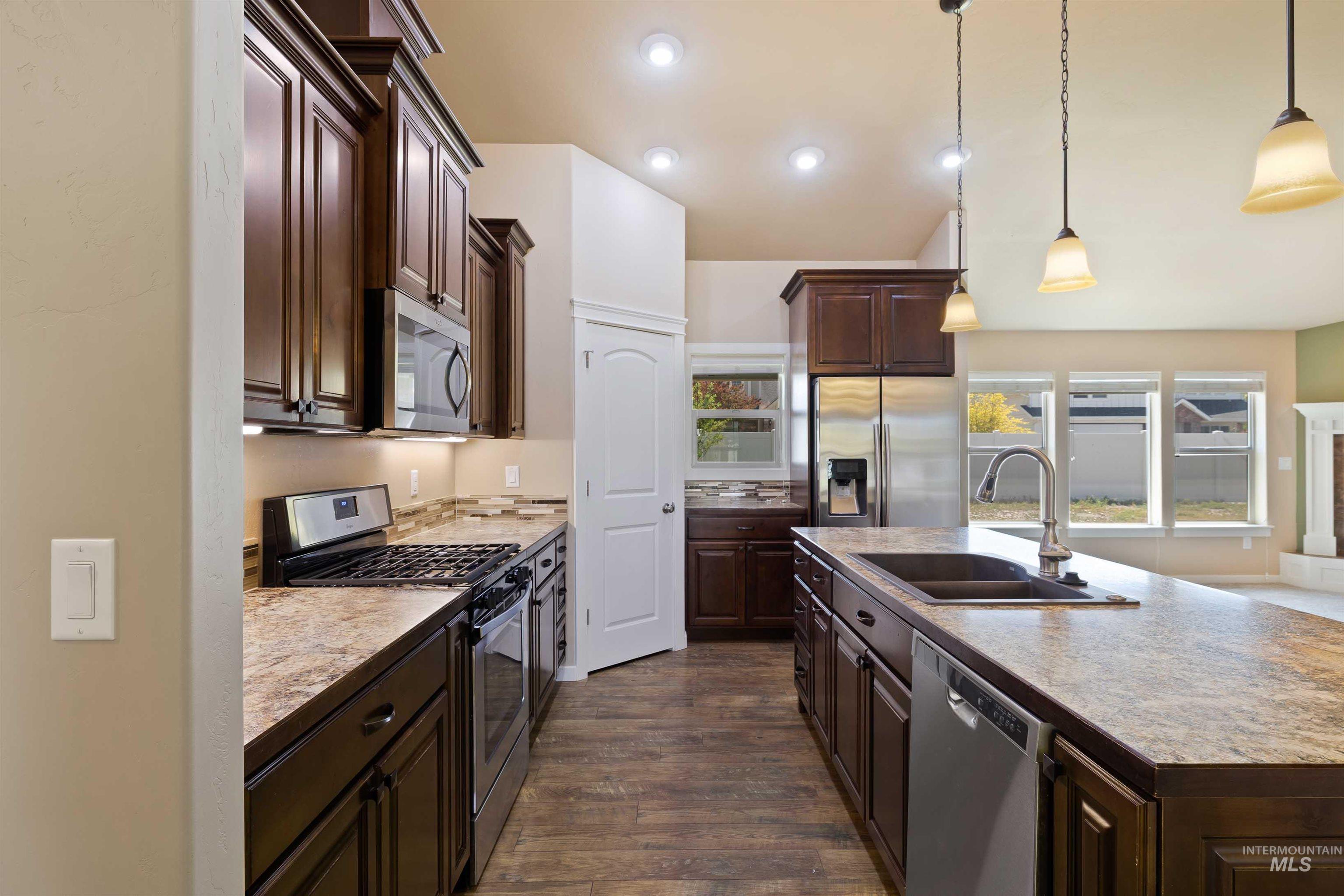 Kitchen featuring appliances with stainless steel finishes, dark brown cabinets, dark wood-type flooring, a kitchen island with sink, and hanging light fixtures