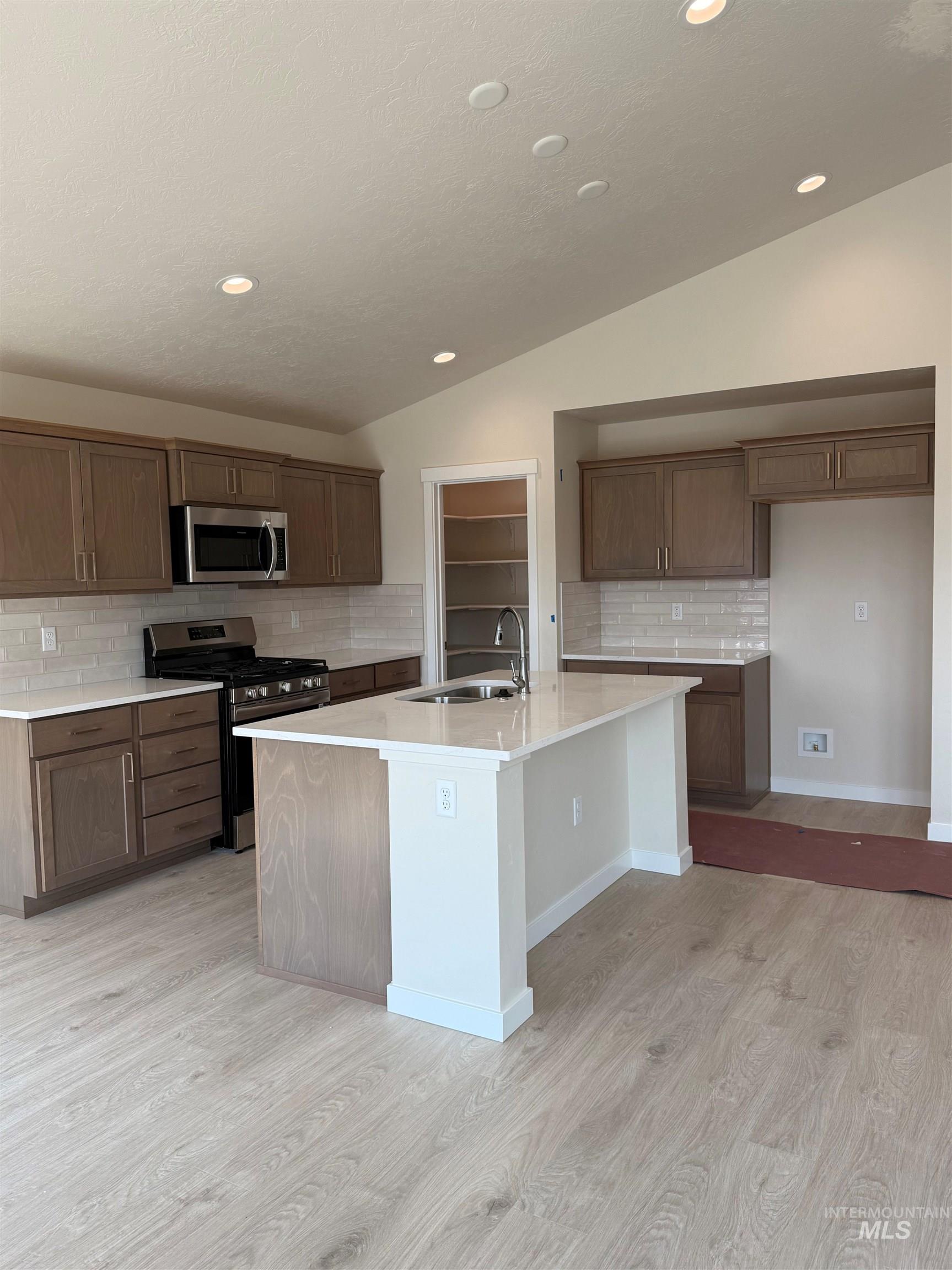 Kitchen featuring decorative backsplash, lofted ceiling, appliances with stainless steel finishes, a kitchen island with sink, and light wood-style flooring