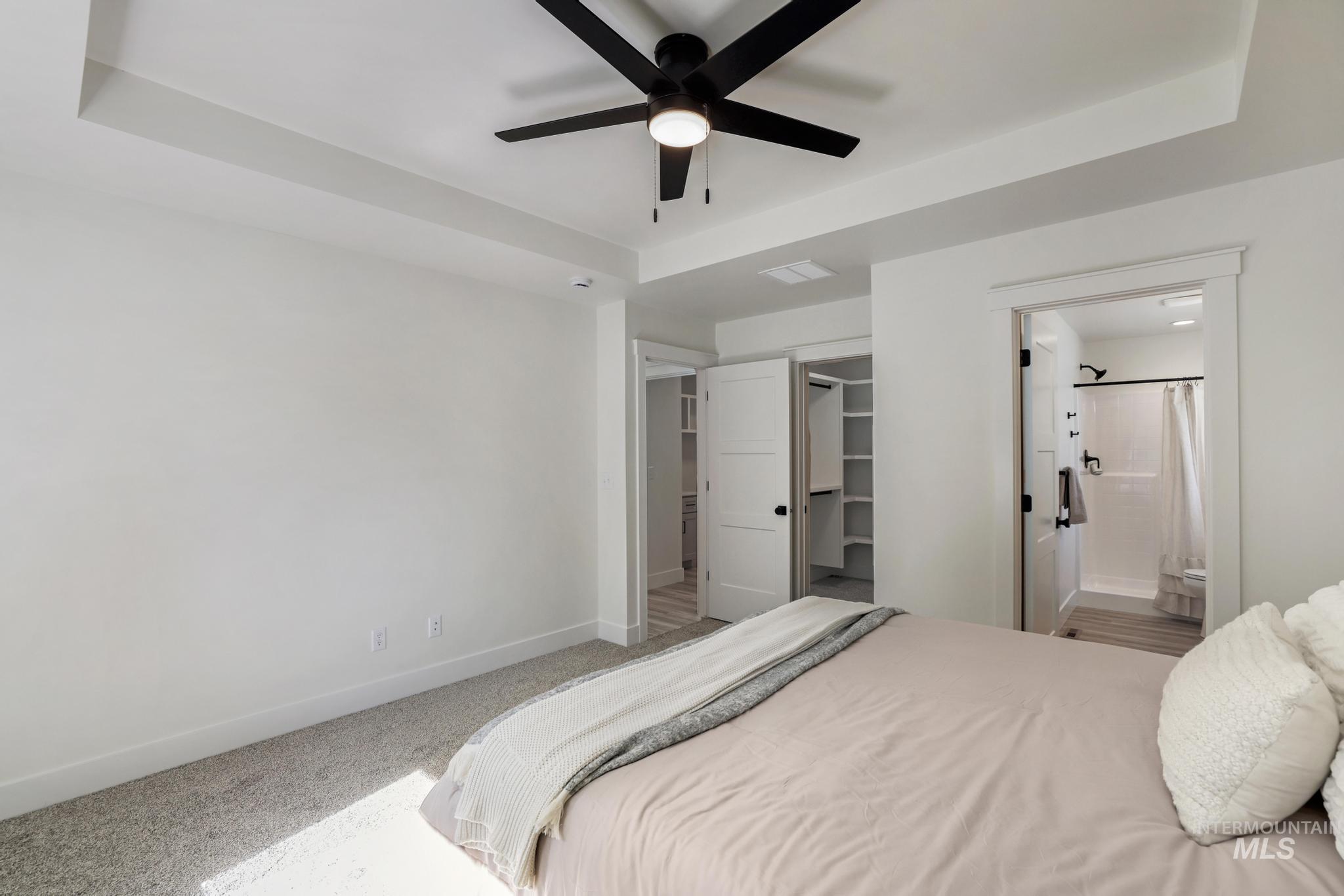 Carpeted bedroom with a tray ceiling, a walk in closet, ensuite bath, and ceiling fan