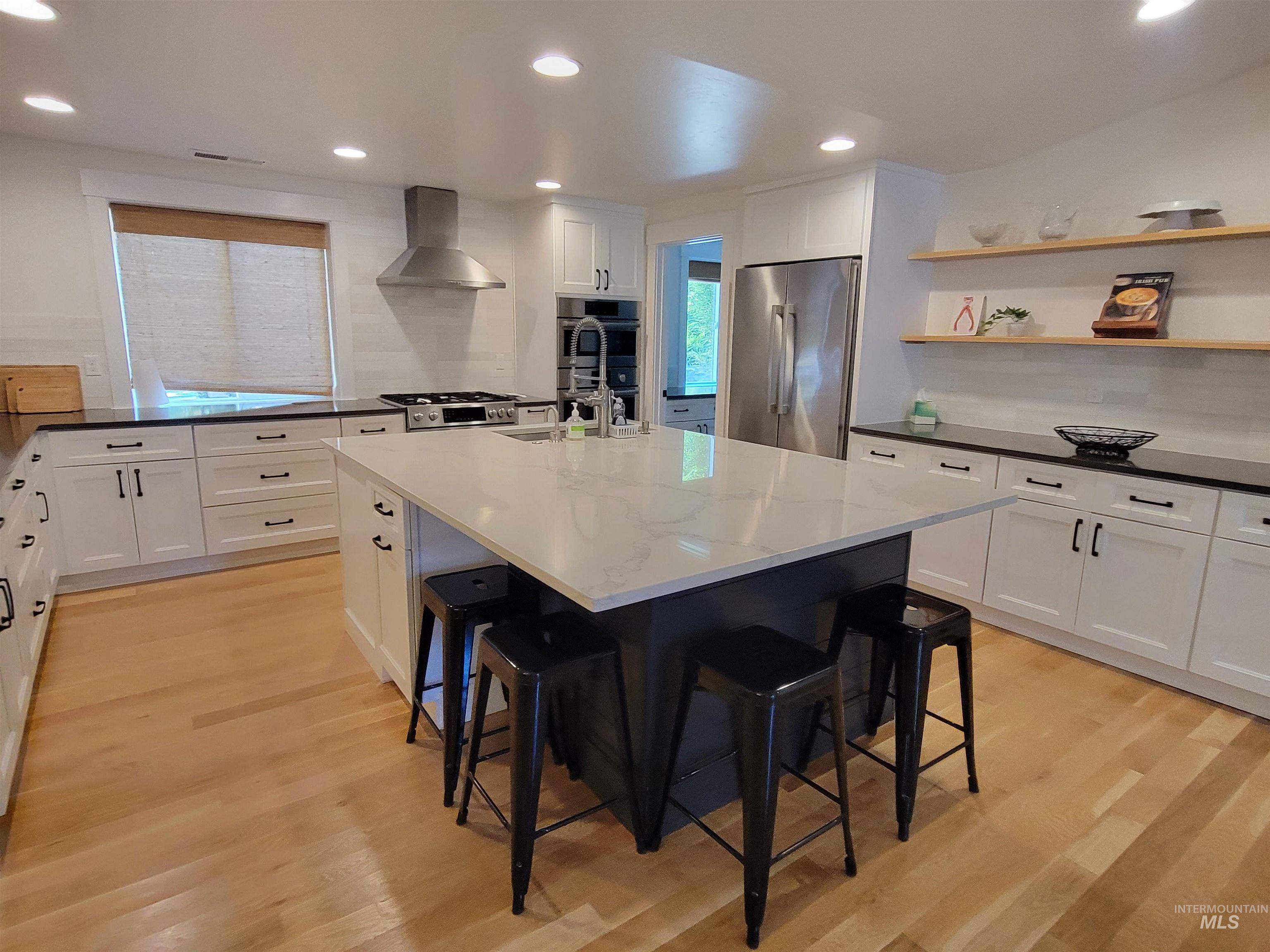 Kitchen with stainless steel appliances, light wood-style flooring, a kitchen bar, wall chimney range hood, and white cabinetry