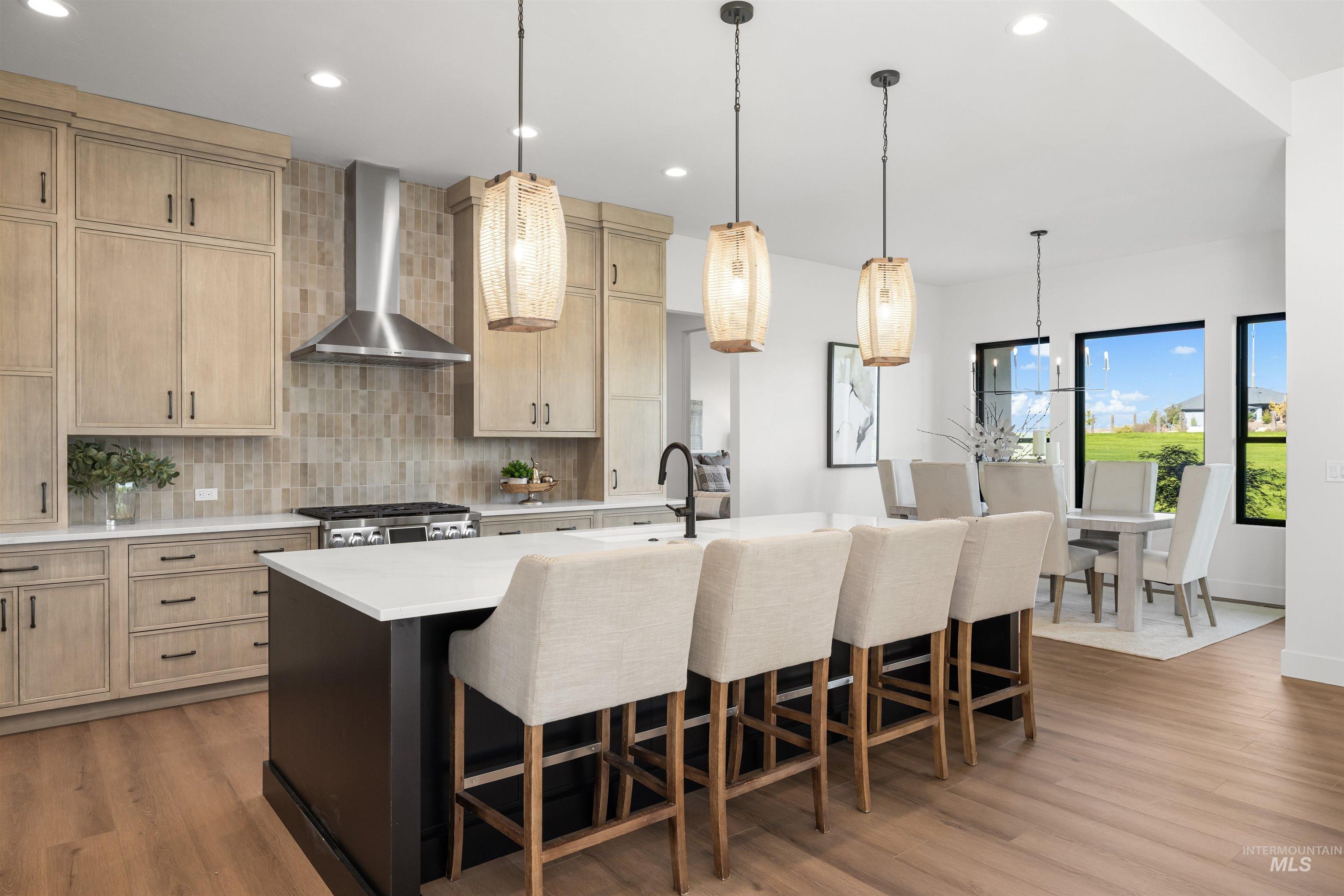 Kitchen featuring light brown cabinetry, backsplash, wall chimney range hood, decorative light fixtures, and a kitchen breakfast bar