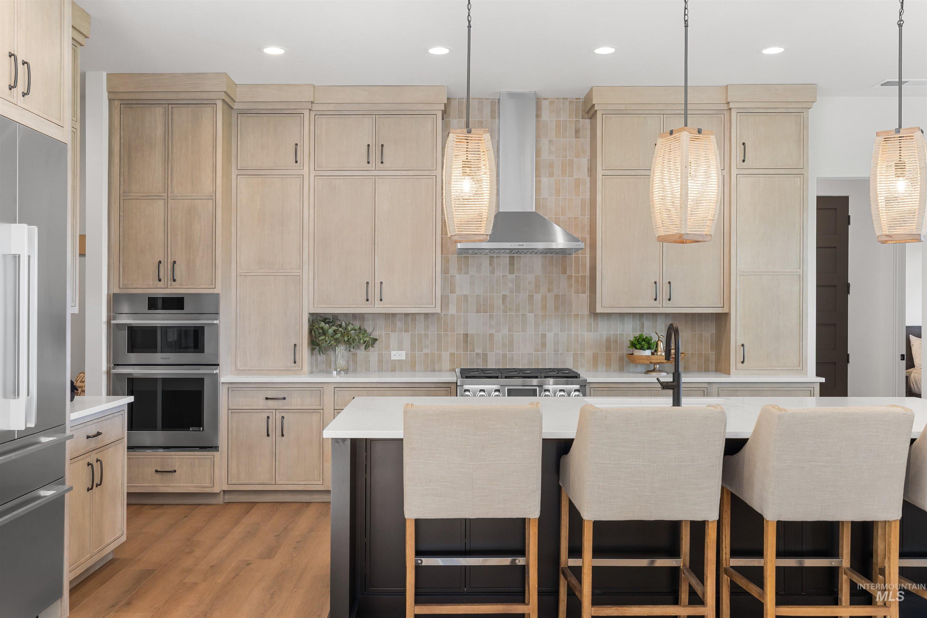 Kitchen with light brown cabinets, pendant lighting, wall chimney range hood, appliances with stainless steel finishes, and recessed lighting