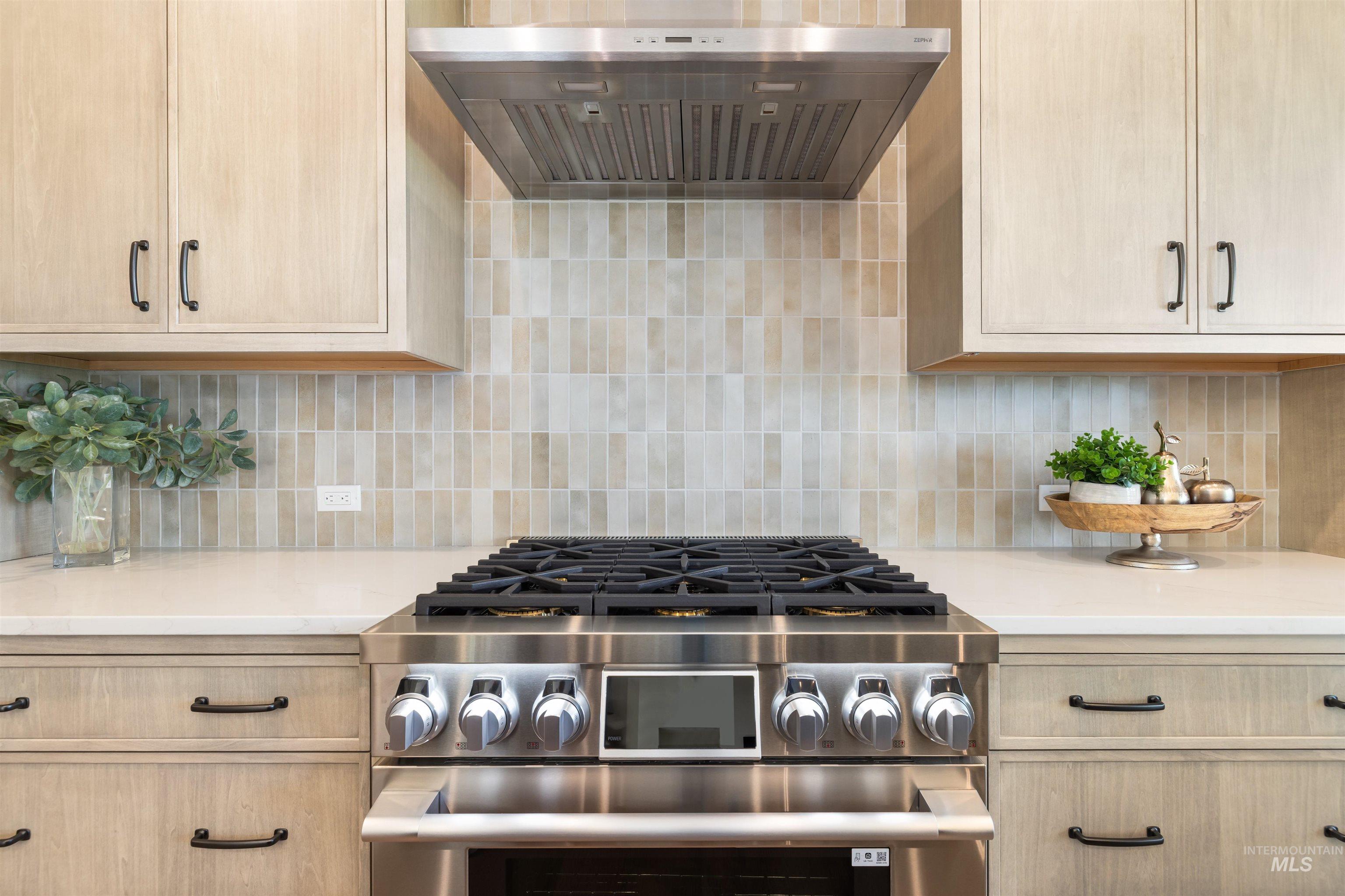 Kitchen with light brown cabinetry, stainless steel range with gas stovetop, ventilation hood, and backsplash