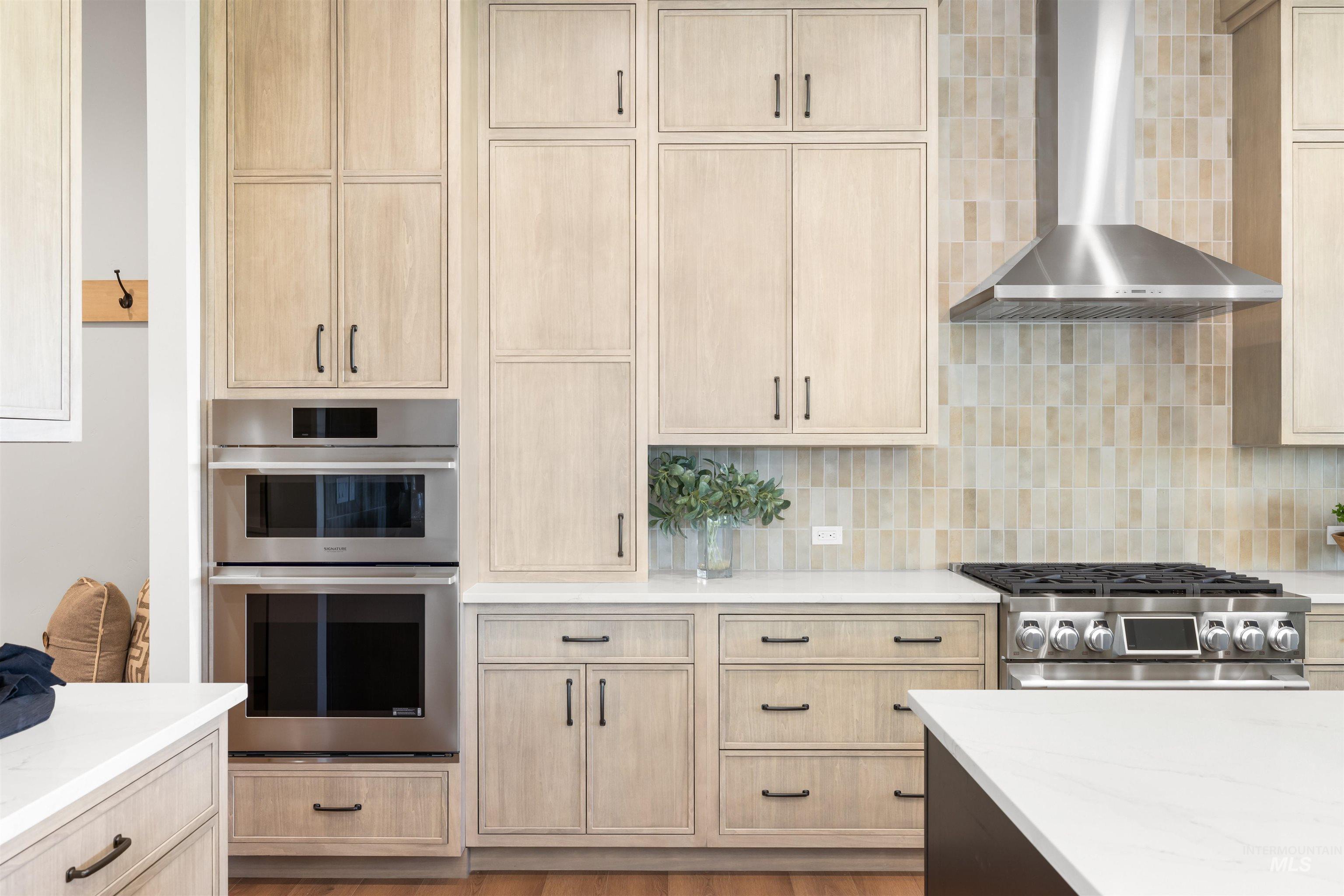 Kitchen with light brown cabinets, wall chimney range hood, appliances with stainless steel finishes, light stone countertops, and tasteful backsplash