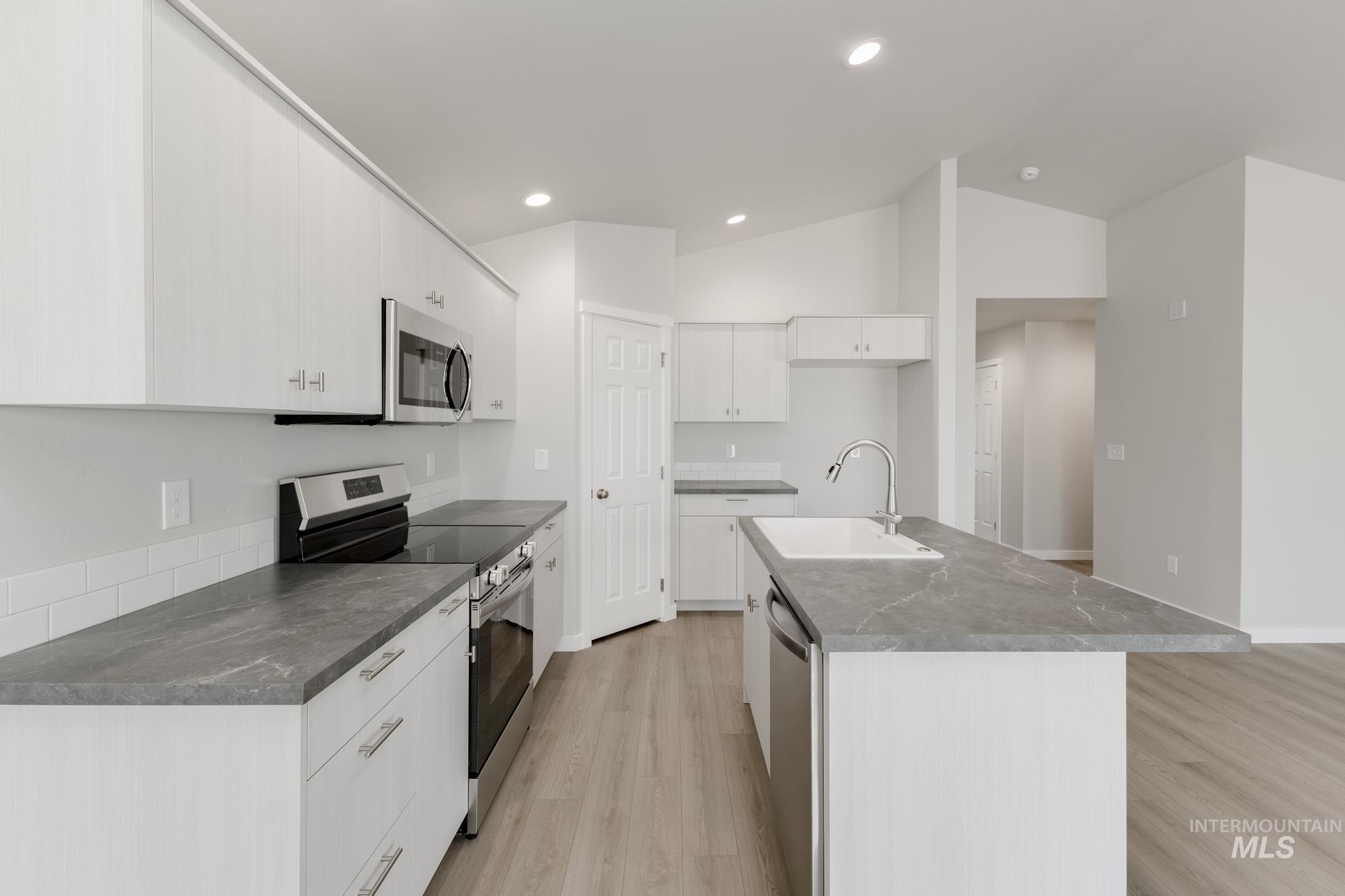 Kitchen featuring stainless steel appliances, vaulted ceiling, white cabinets, light wood-style flooring, and a kitchen island with sink