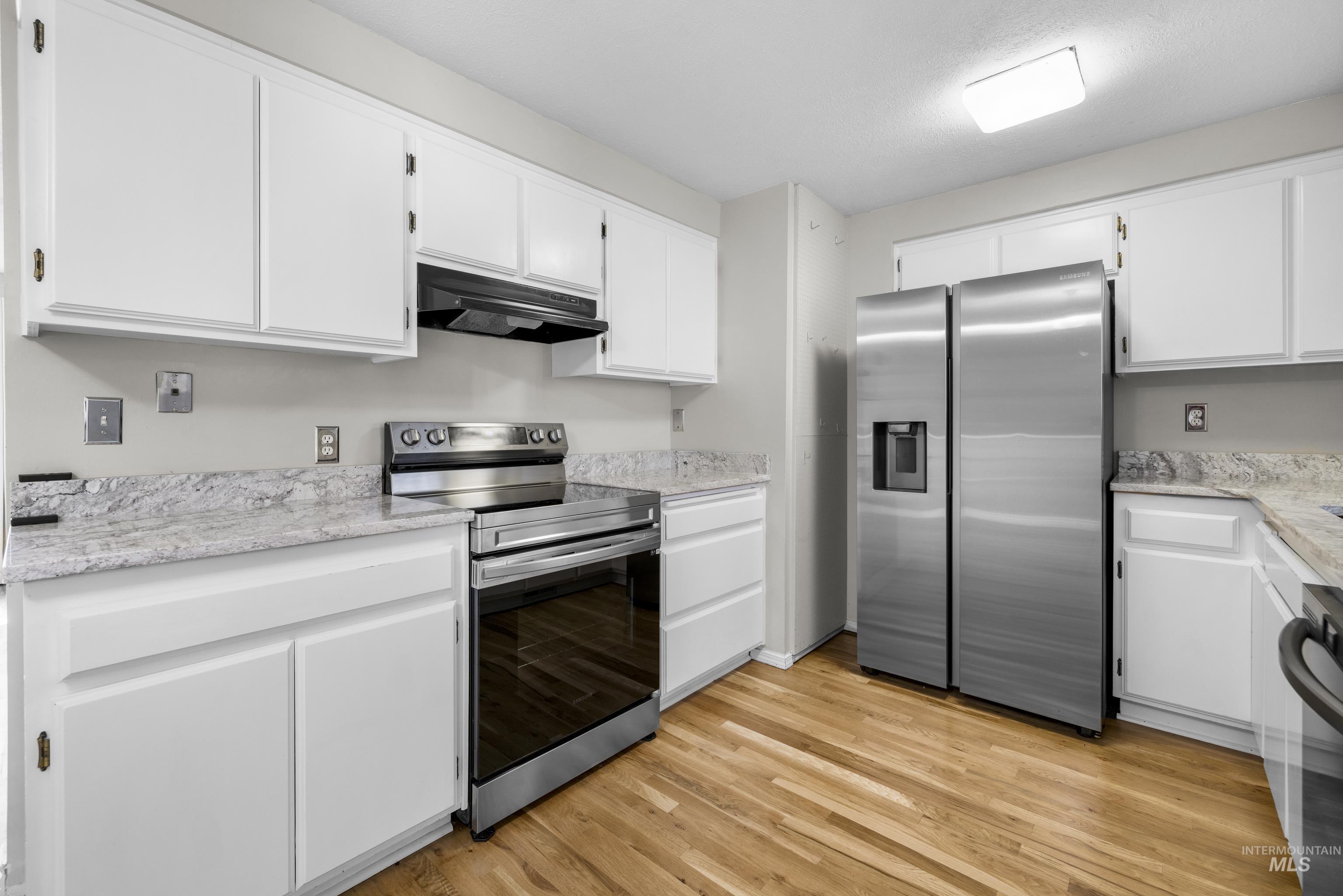 Kitchen with appliances with stainless steel finishes, under cabinet range hood, white cabinetry, light wood-type flooring, and light stone countertops