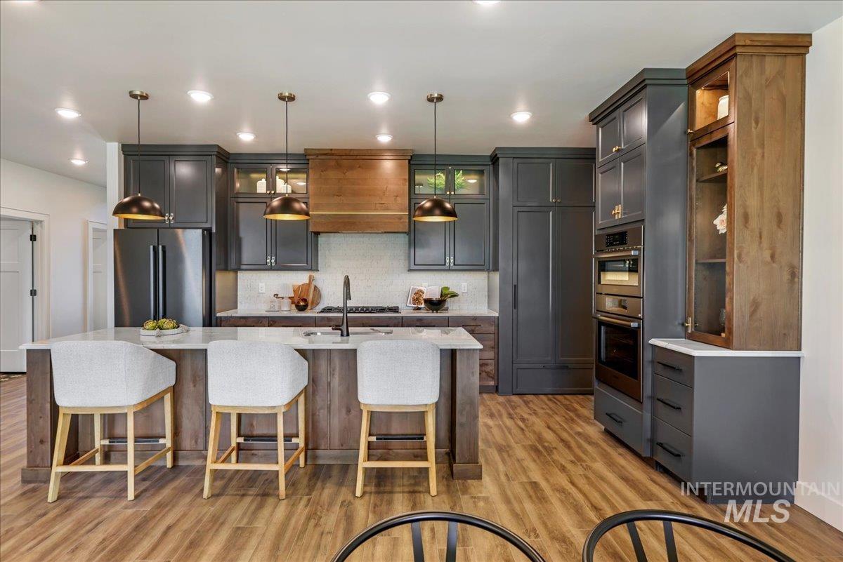 Kitchen with stainless steel appliances, light wood-style flooring, backsplash, an island with sink, and pendant lighting