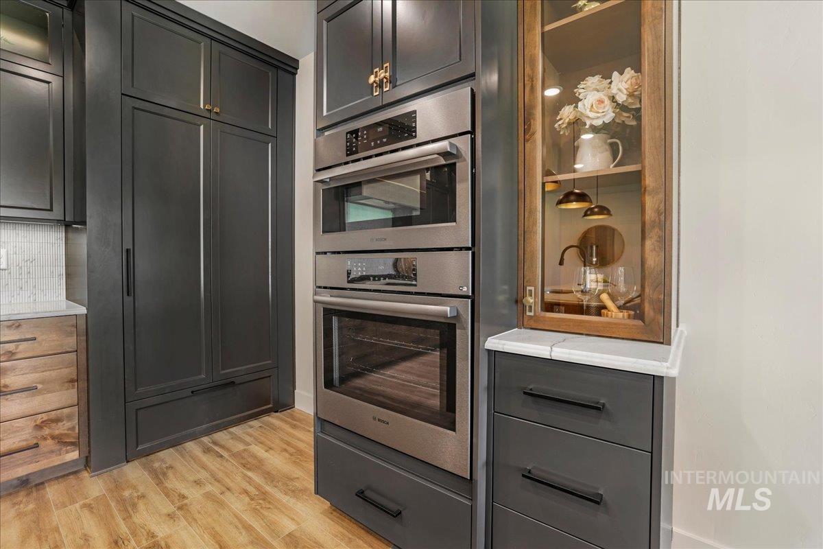Kitchen with double oven, light wood finished floors, decorative backsplash, light countertops, and glass insert cabinets