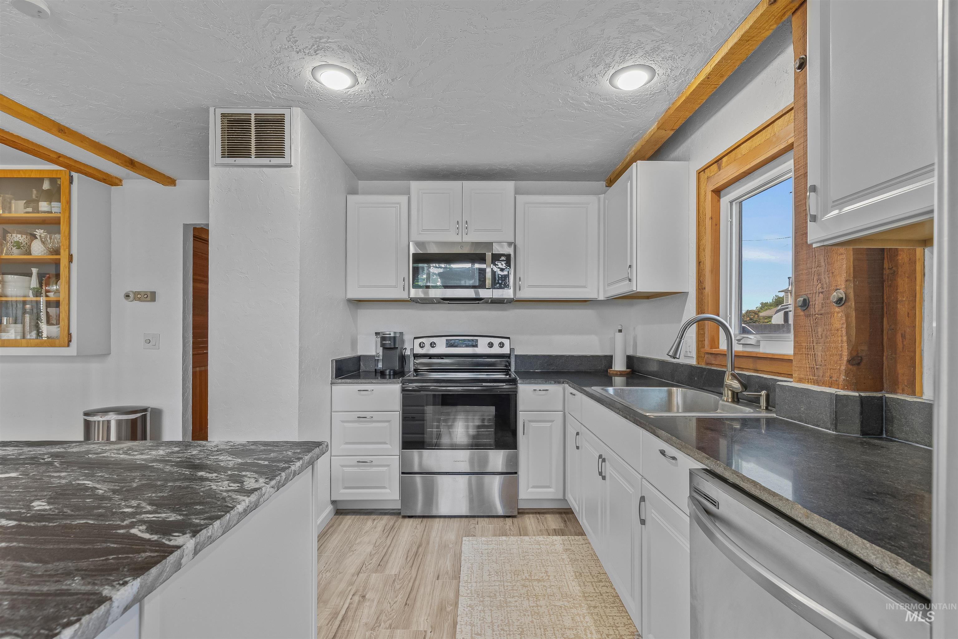 Kitchen with appliances with stainless steel finishes, a textured ceiling, white cabinetry, light wood-style flooring, and dark stone countertops