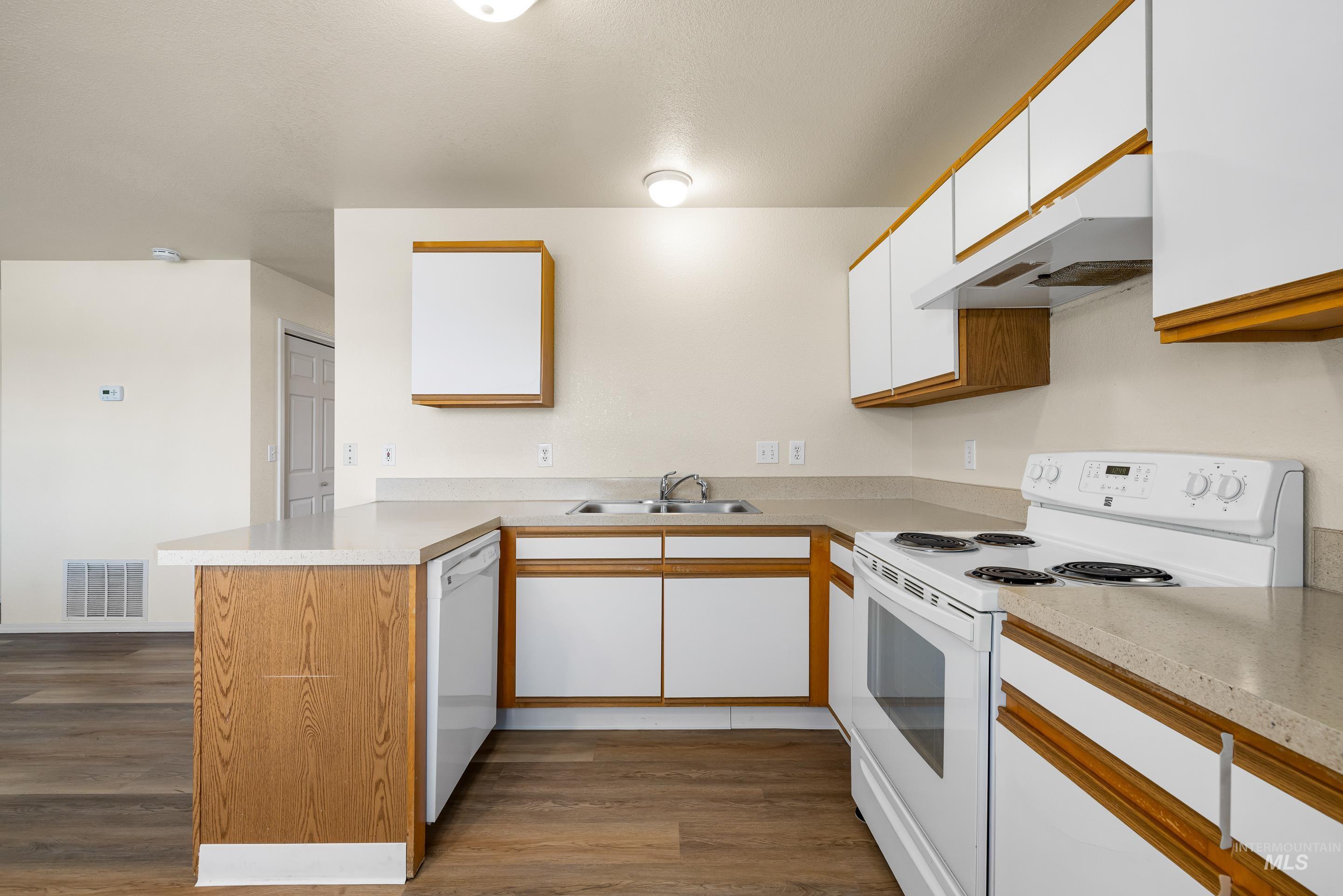 Kitchen with white appliances, a peninsula, under cabinet range hood, white cabinets, and dark wood-style floors