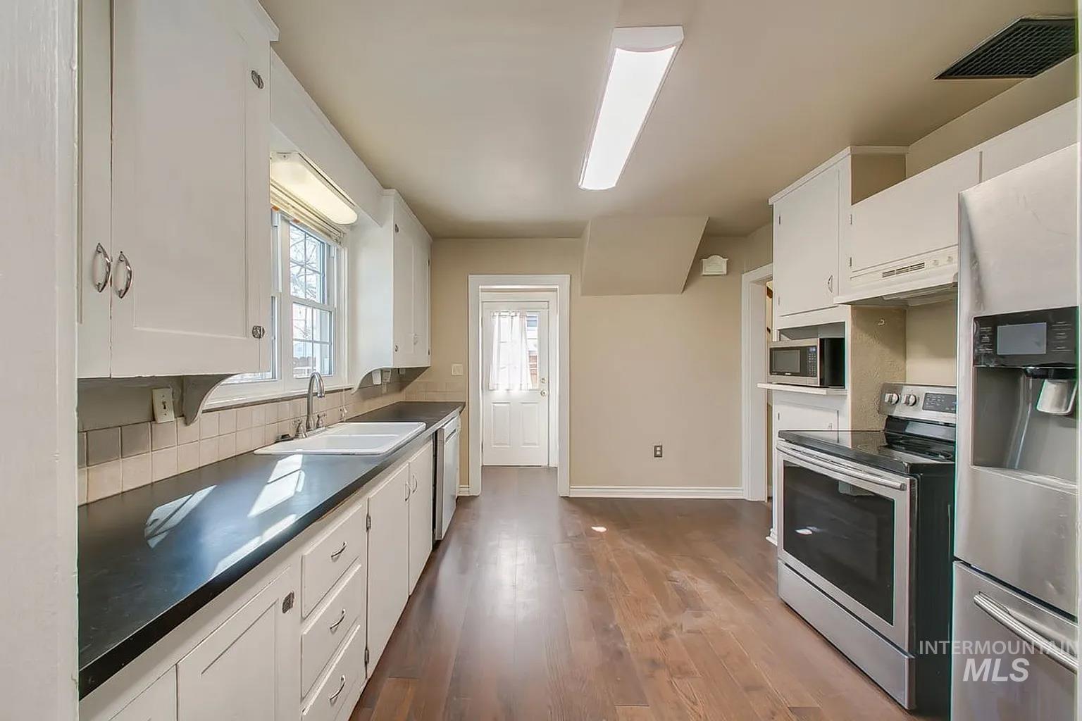 Kitchen featuring appliances with stainless steel finishes, backsplash, white cabinets, dark wood-style floors, and dark countertops