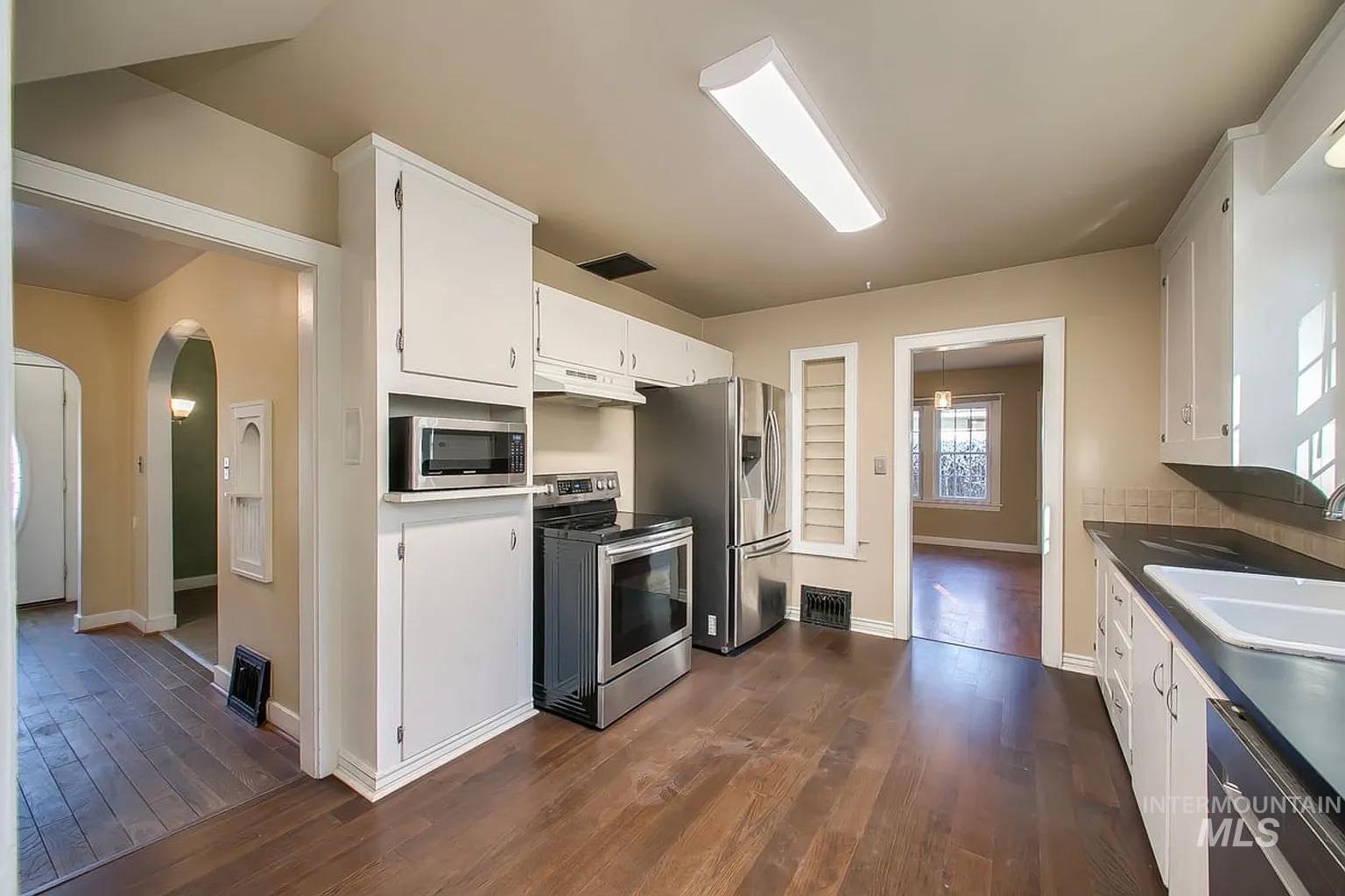 Kitchen with stainless steel appliances, under cabinet range hood, arched walkways, white cabinetry, and dark wood-type flooring