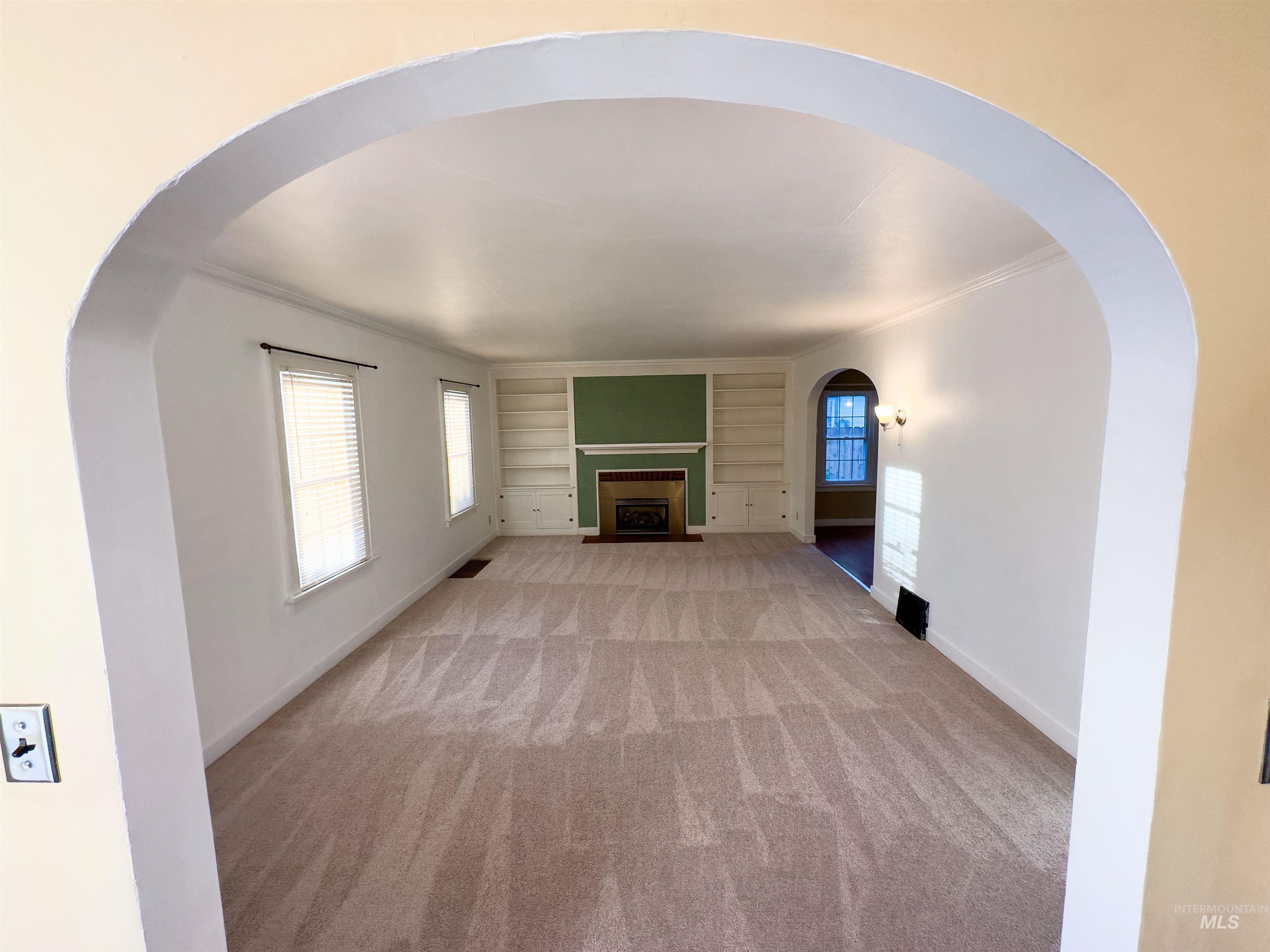 Unfurnished living room with light carpet, a fireplace with flush hearth, built in features, crown molding, and arched walkways