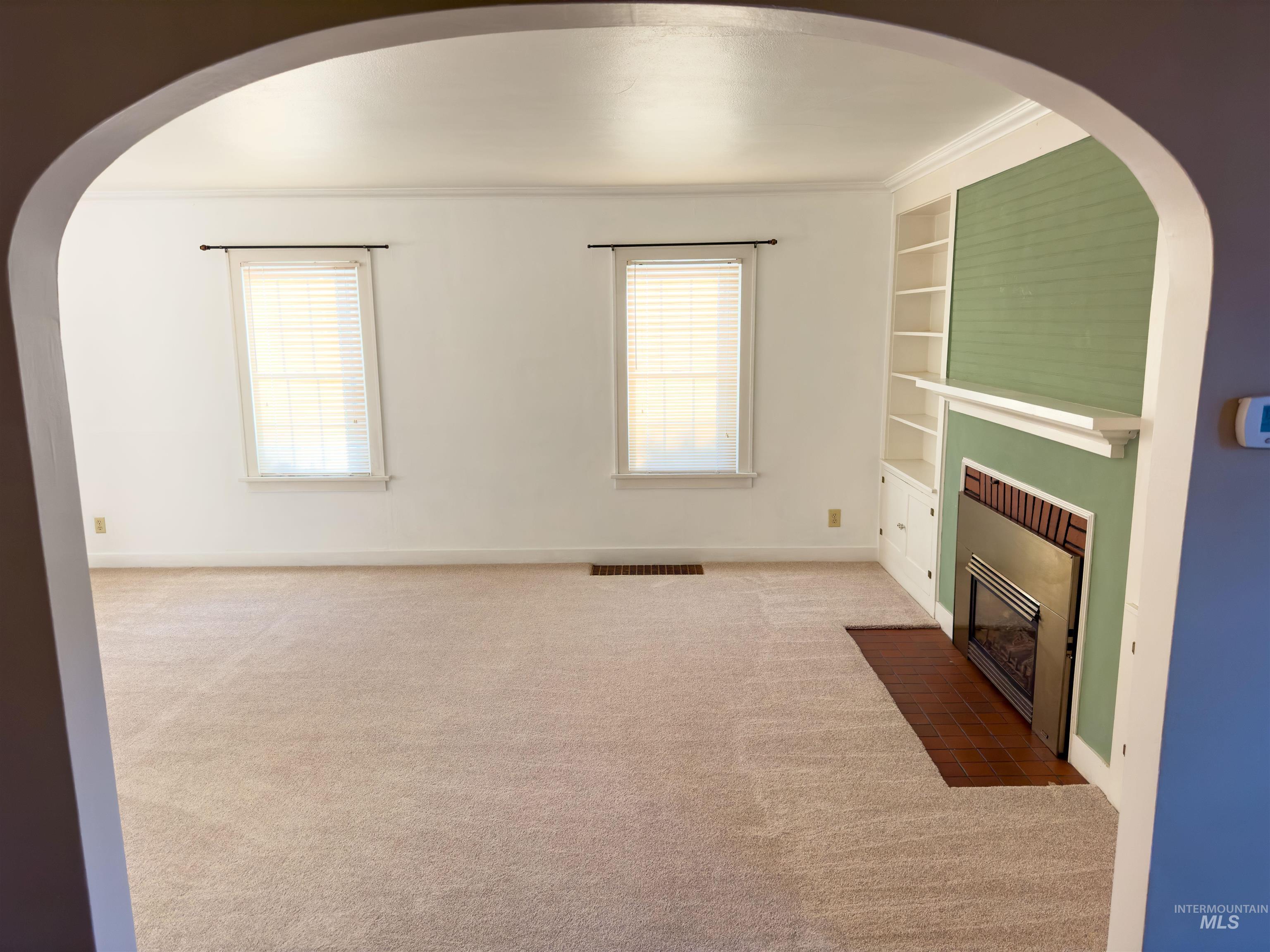 Unfurnished living room featuring built in shelves, carpet, plenty of natural light, a fireplace with flush hearth, and crown molding