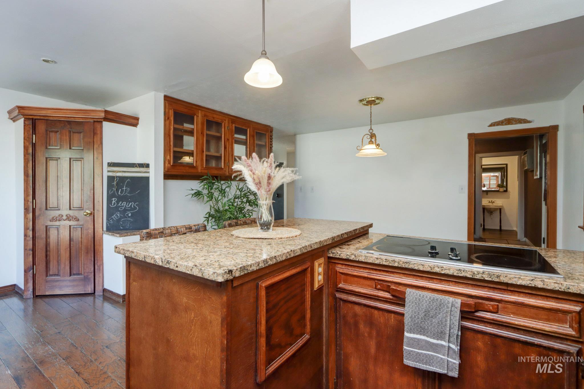 Kitchen featuring black electric cooktop, decorative light fixtures, glass insert cabinets, dark wood-style flooring, and light stone counters