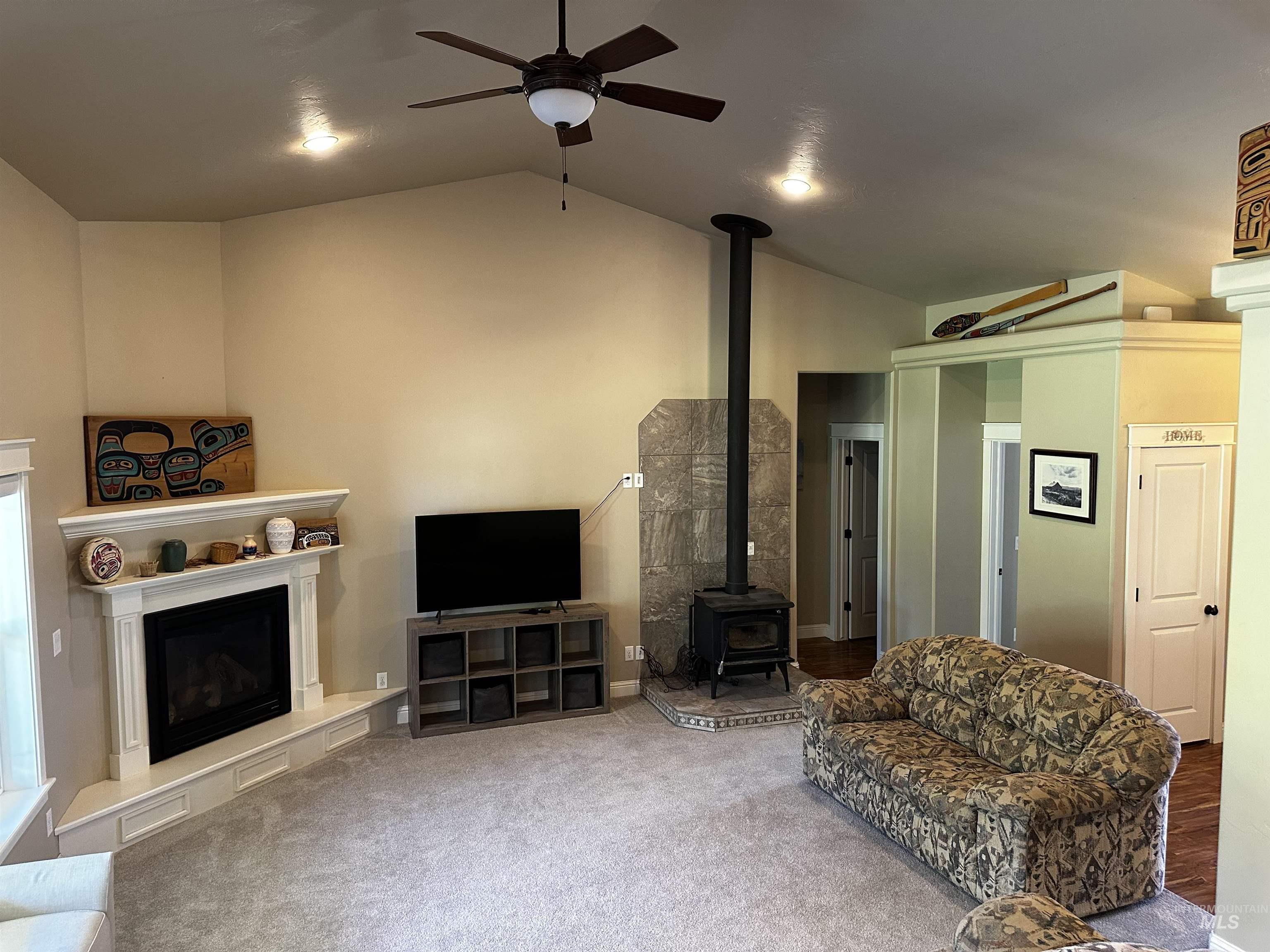 Living room featuring a wood stove, carpet floors, a ceiling fan, high vaulted ceiling, and a fireplace