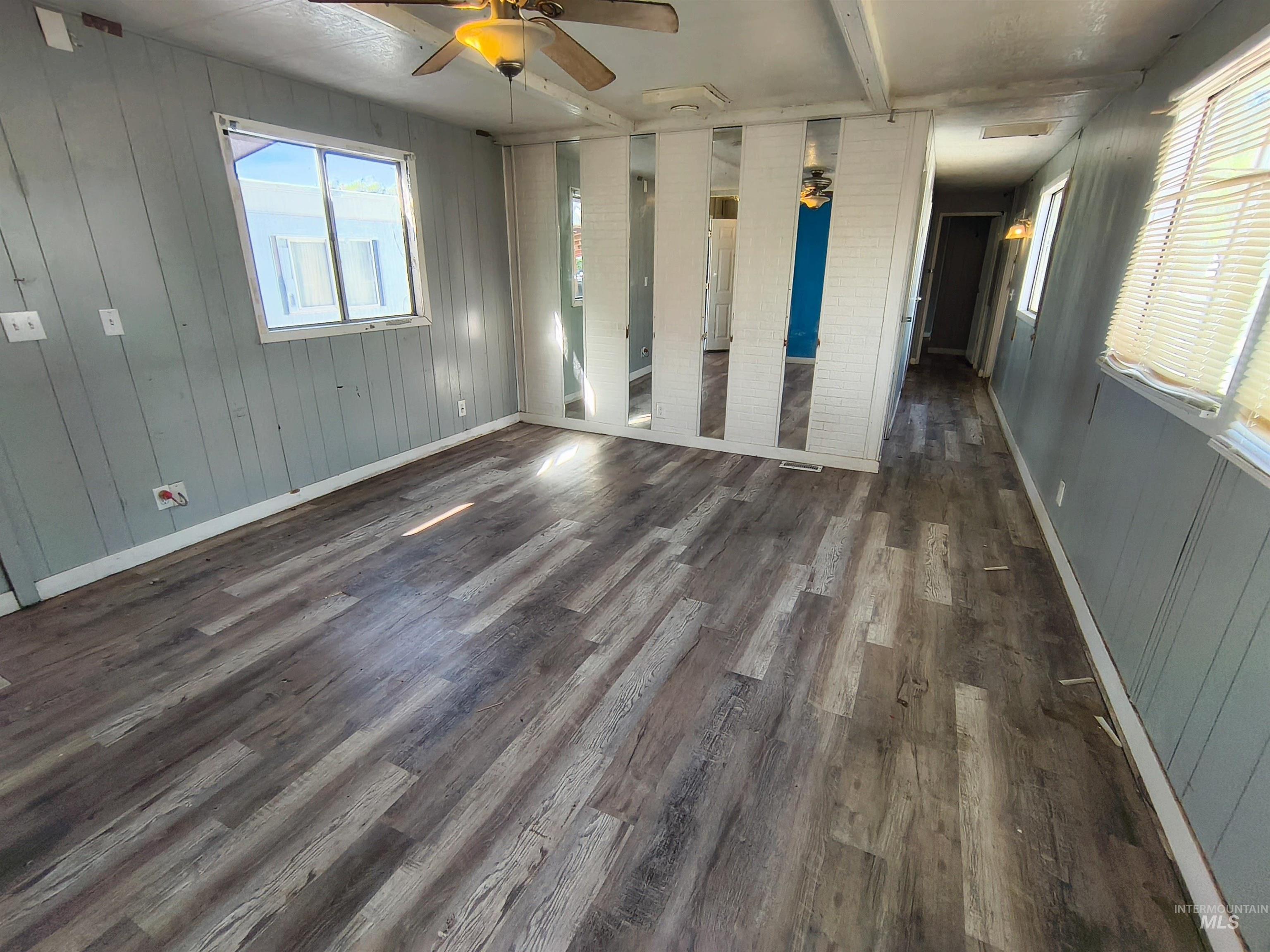 Empty room with ceiling fan, healthy amount of natural light, wooden walls, dark wood-style flooring, and beam ceiling