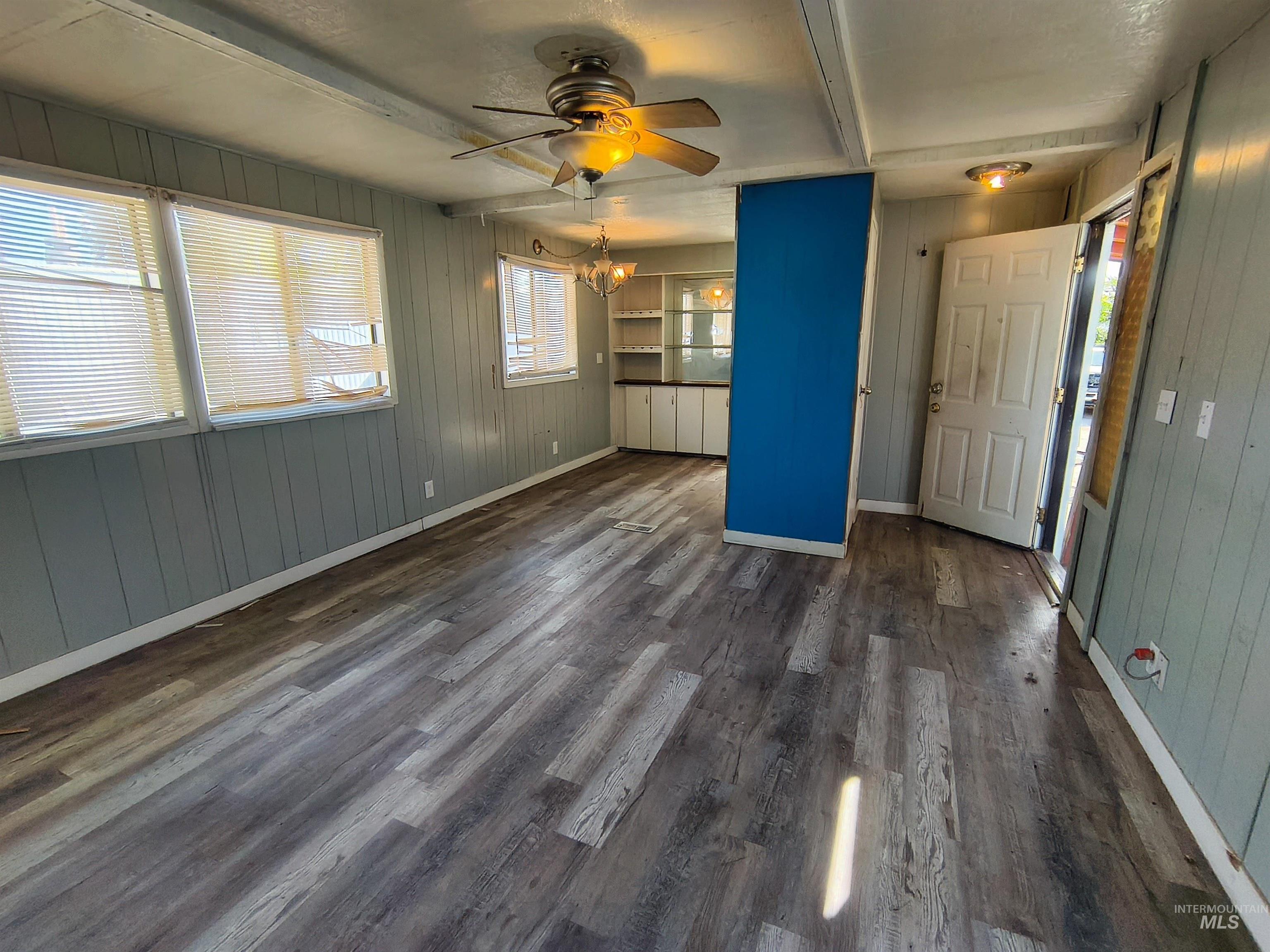 Unfurnished living room with wooden walls, plenty of natural light, beamed ceiling, a ceiling fan, and dark wood-type flooring
