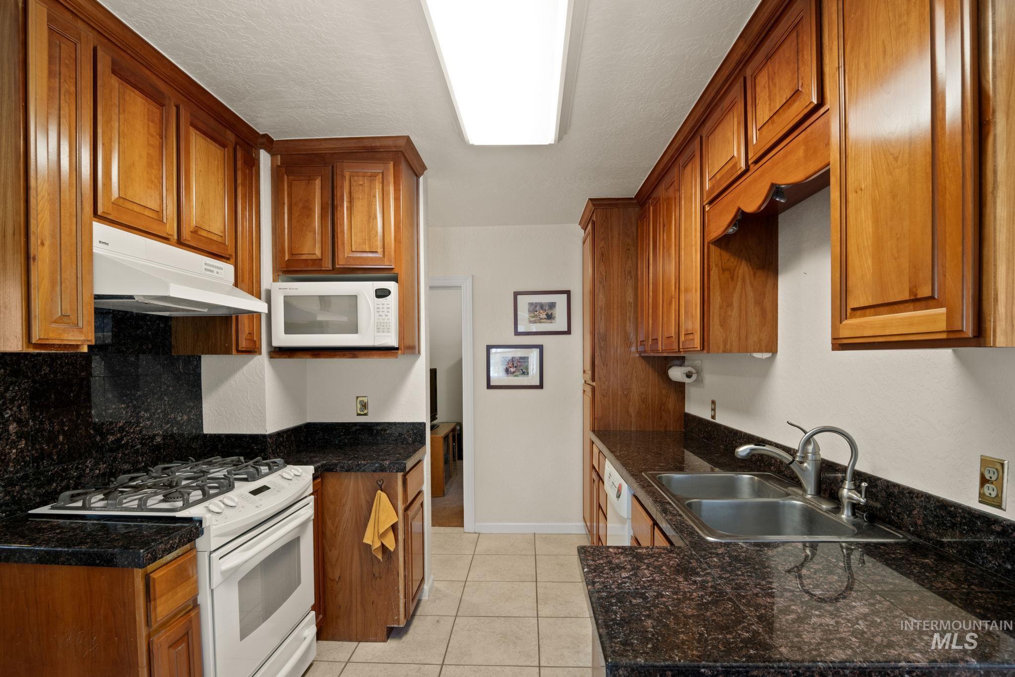 Kitchen with white appliances, brown cabinetry, light tile patterned flooring, under cabinet range hood, and dark countertops