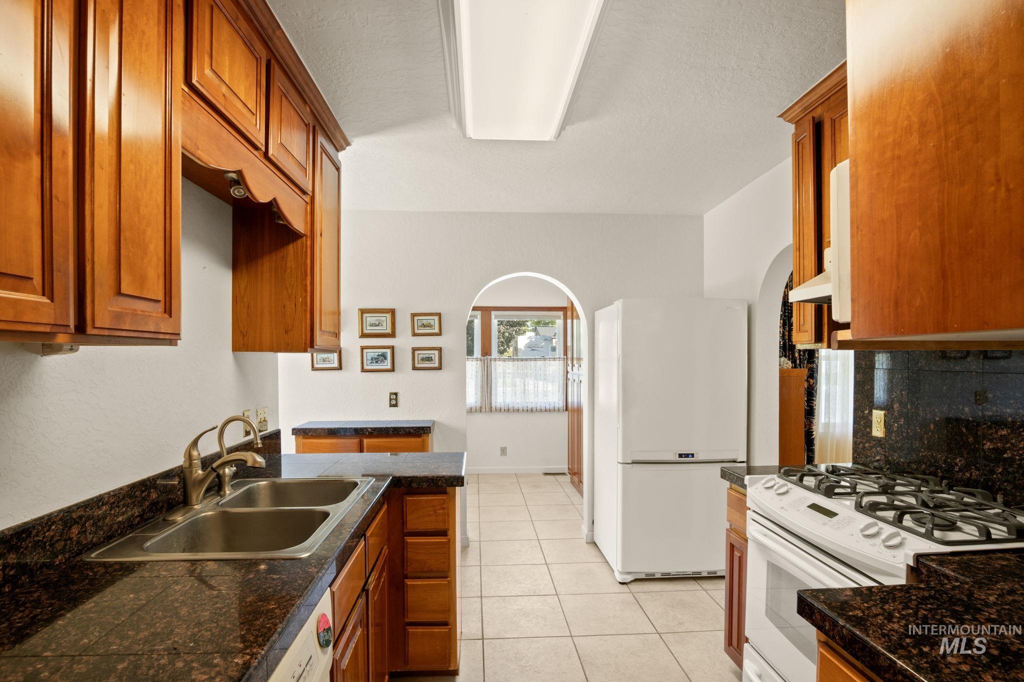 Kitchen with white appliances, light tile patterned floors, arched walkways, brown cabinets, and backsplash