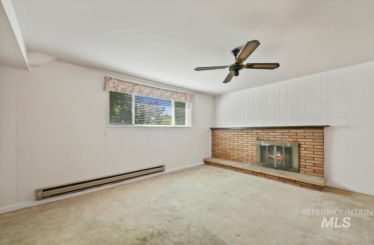 Unfurnished living room featuring baseboard heating, carpet floors, a brick fireplace, and ceiling fan