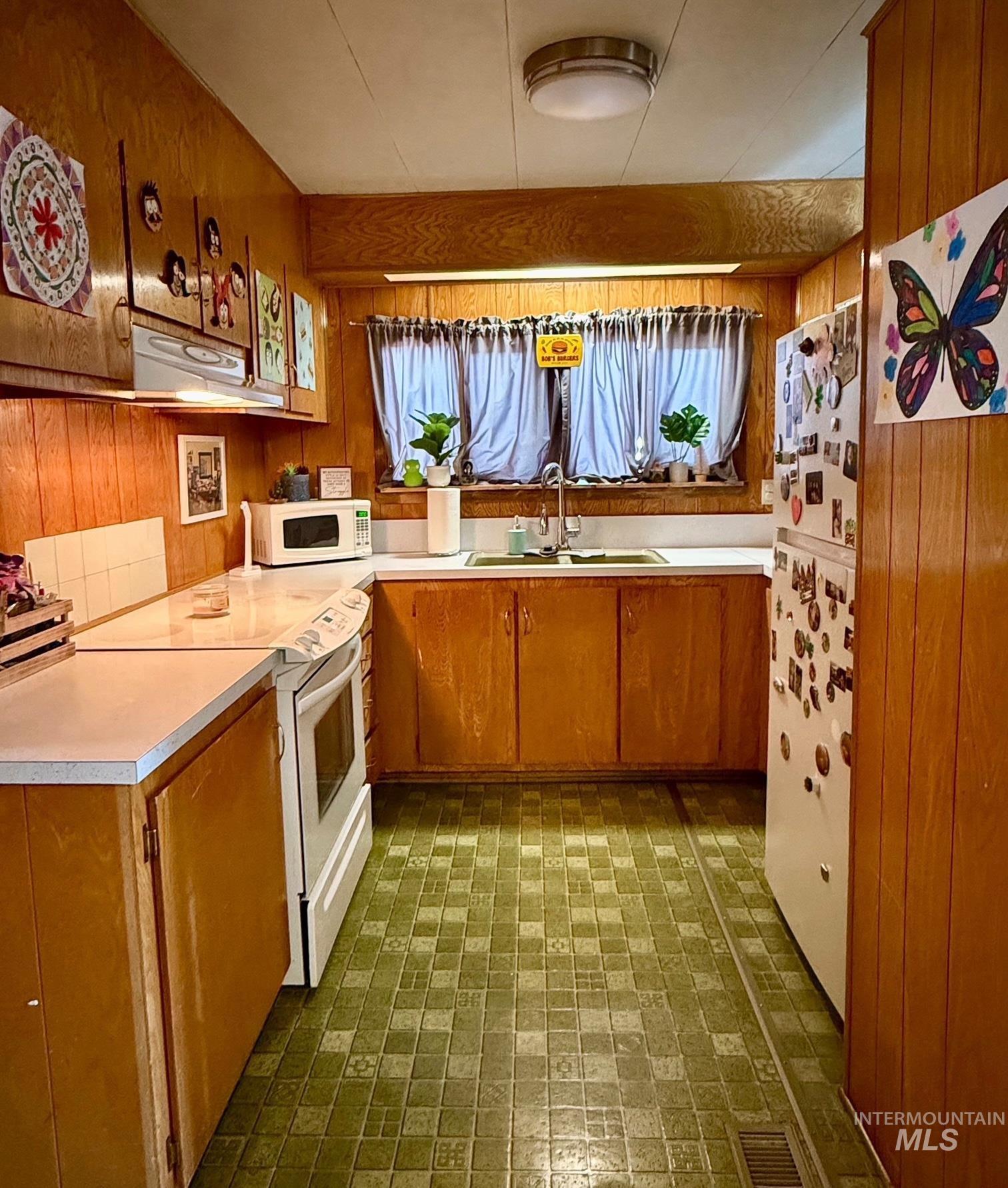 Kitchen with brown cabinetry, wood walls, white appliances, light countertops, and under cabinet range hood
