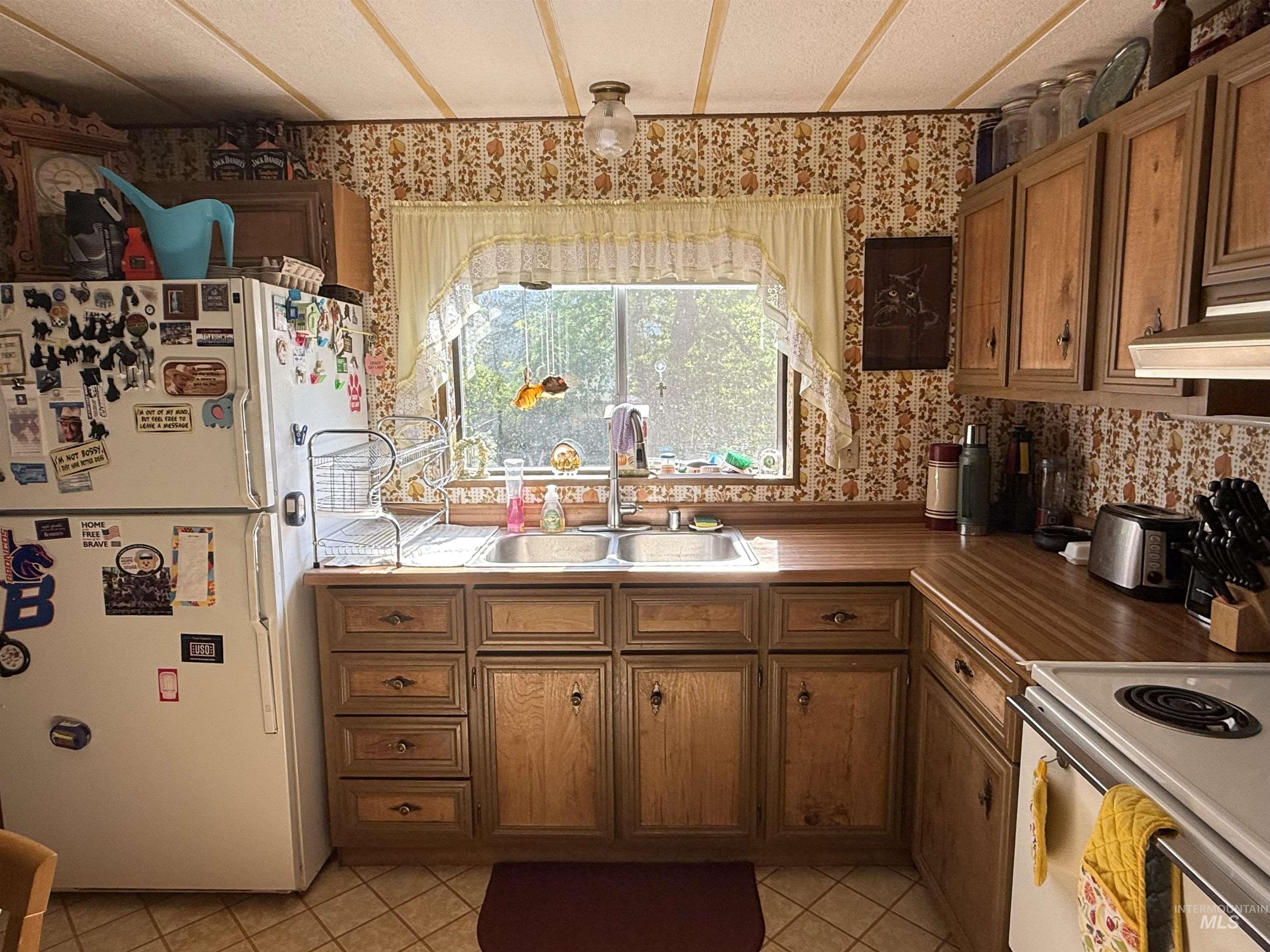 Kitchen with freestanding refrigerator, wallpapered walls, brown cabinets, light tile patterned floors, and range