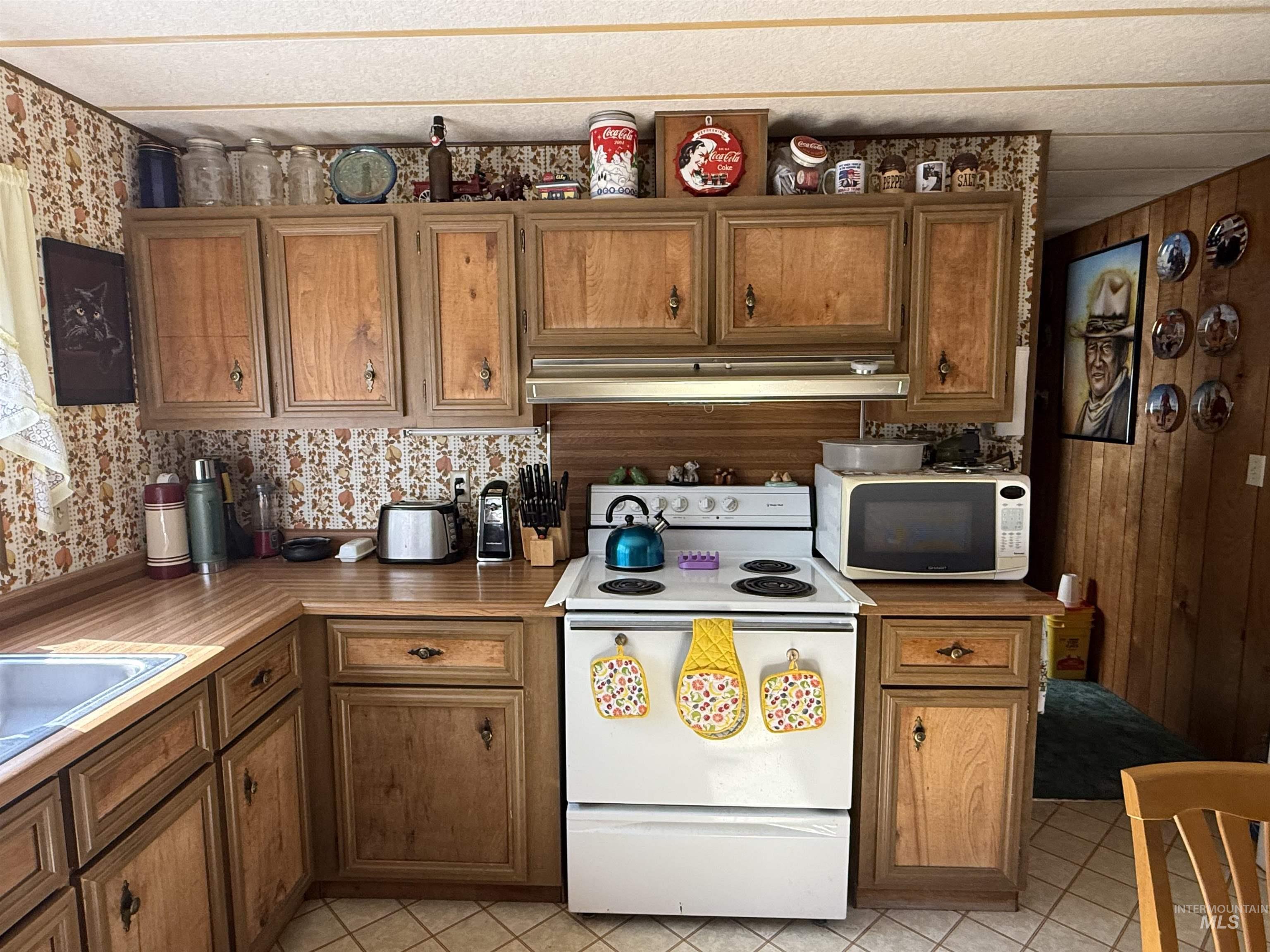 Kitchen featuring white appliances, under cabinet range hood, brown cabinetry, light tile patterned floors, and wallpapered walls