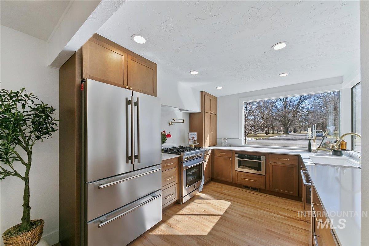 Kitchen with brown cabinetry, high quality appliances, light wood finished floors, recessed lighting, and light stone counters