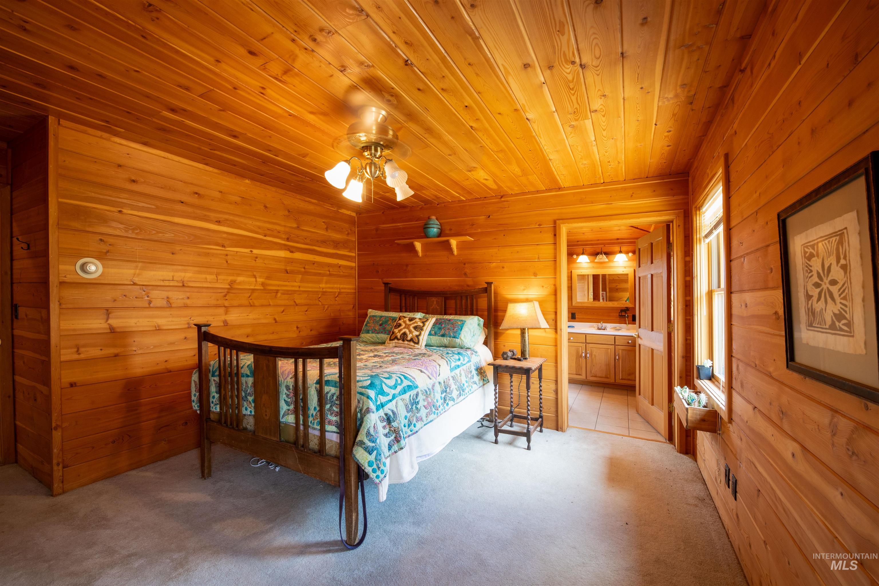 Bedroom with wood walls, light colored carpet, wood ceiling, connected bathroom, and a ceiling fan