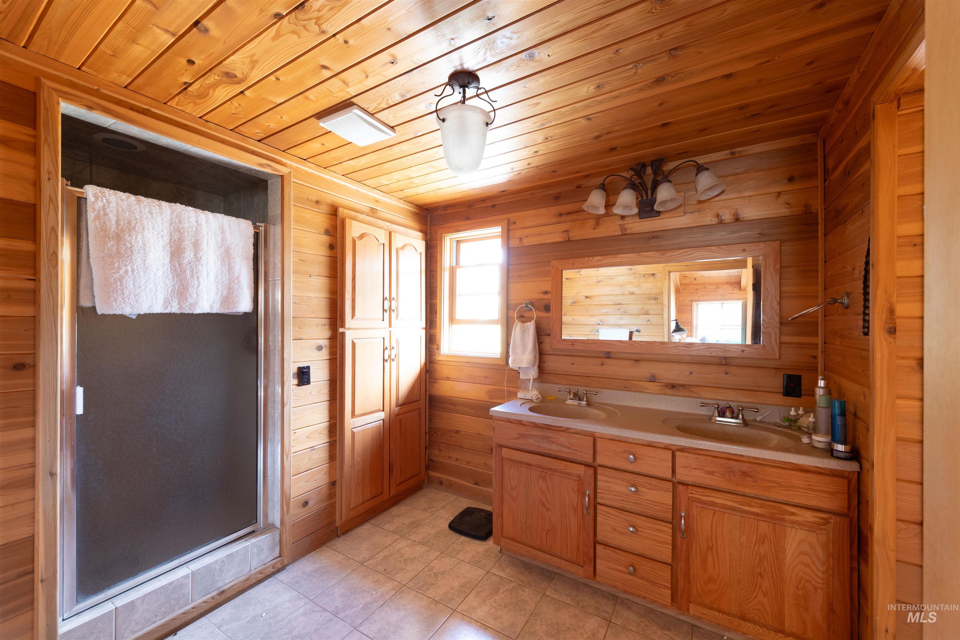 Bathroom featuring a shower stall, wood walls, double vanity, wooden ceiling, and light tile patterned floors