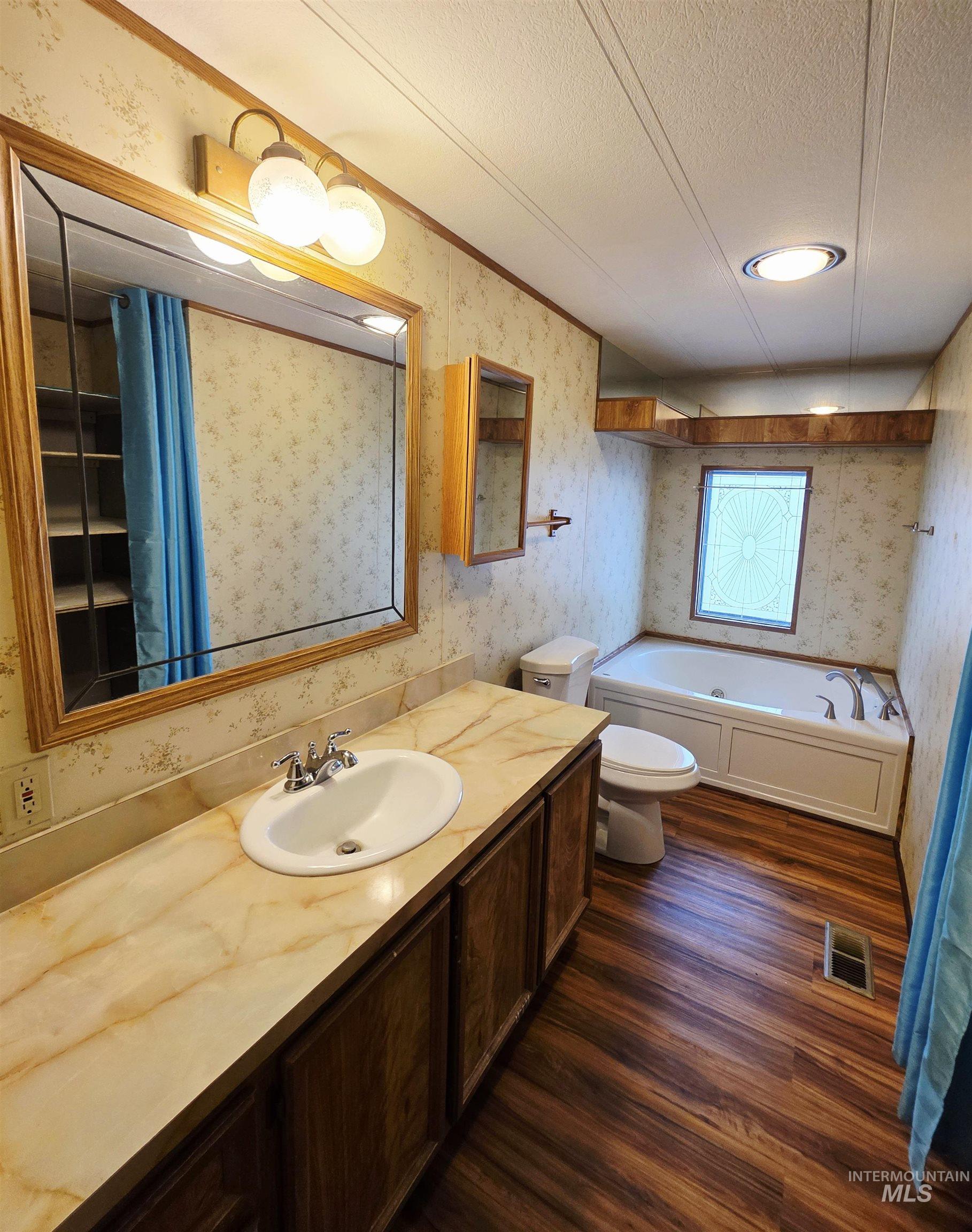 Full bathroom featuring wallpapered walls, dark wood-type flooring, a garden tub, vanity, and a textured ceiling