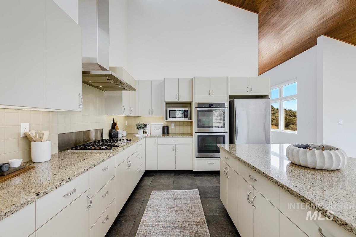 Kitchen with wall chimney range hood, stainless steel appliances, decorative backsplash, light stone countertops, and white cabinets