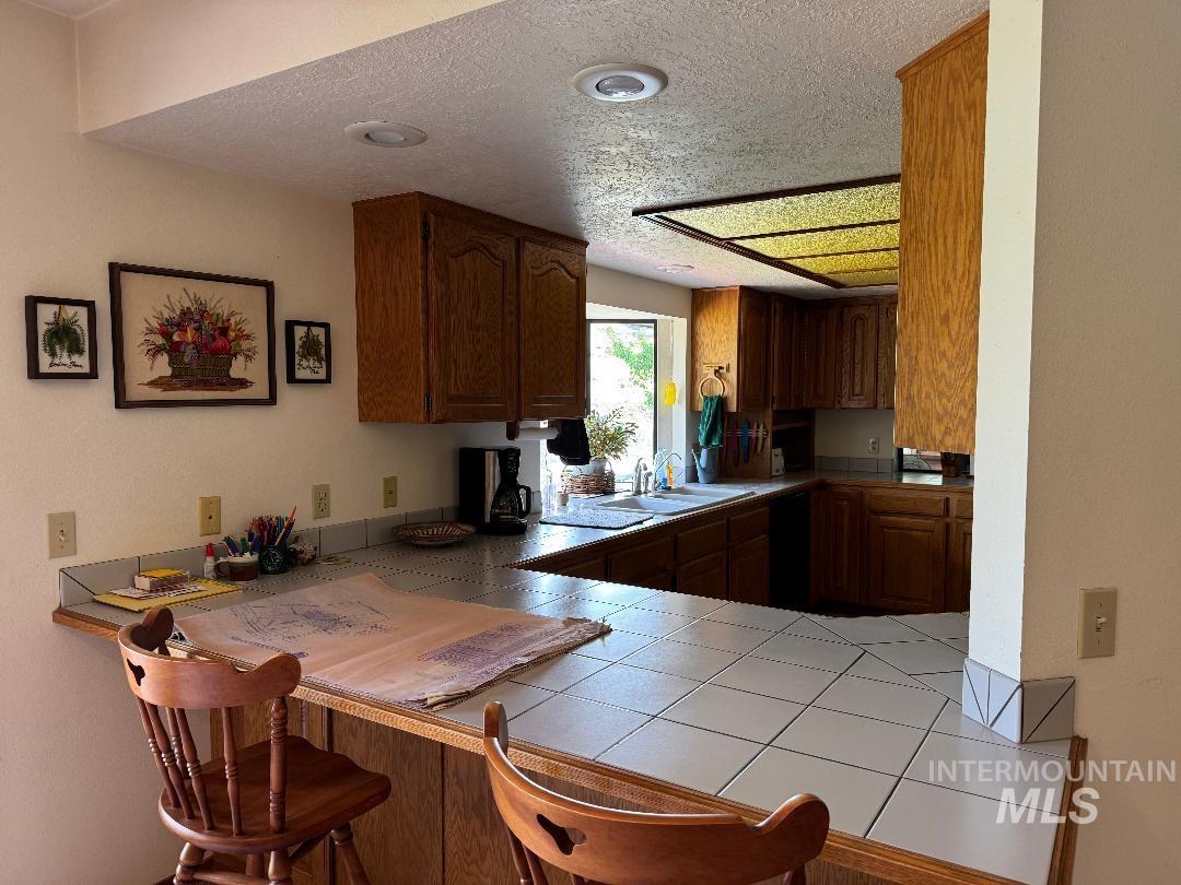 Kitchen with tile counters, brown cabinetry, a textured ceiling, a breakfast bar, and dishwasher