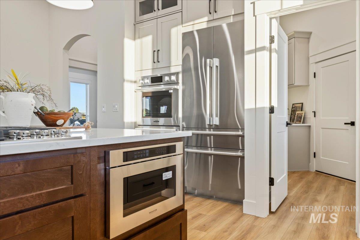 Kitchen with stainless steel appliances, light wood-type flooring, light stone counters, glass insert cabinets, and dark brown cabinets