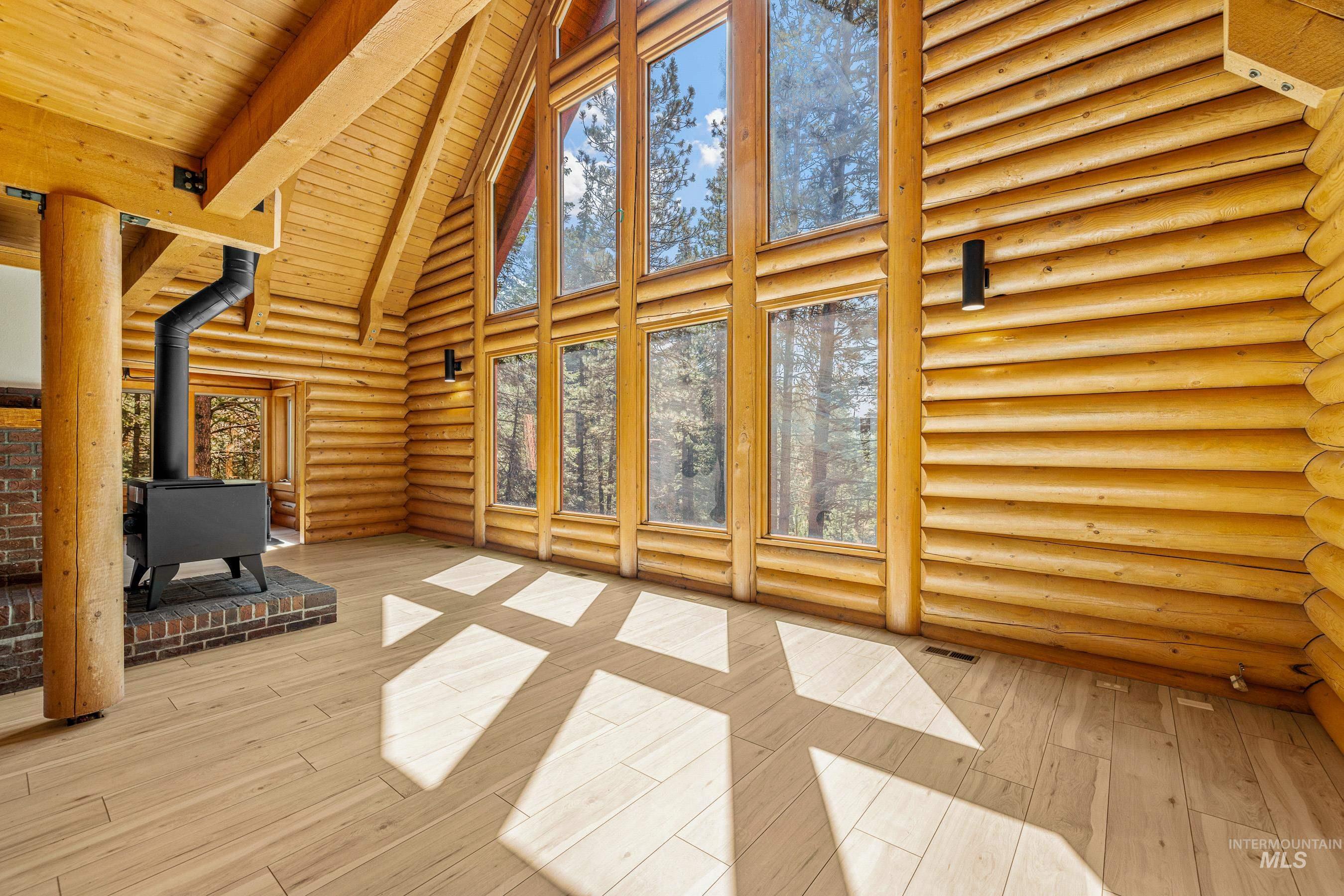 Unfurnished living room with a wood stove, rustic walls, high vaulted ceiling, light wood-style floors, and a wooden ceiling with exposed beams