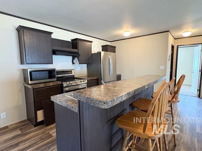 Kitchen with dark brown cabinets, stainless steel appliances, a kitchen breakfast bar, dark wood-style floors, and crown molding