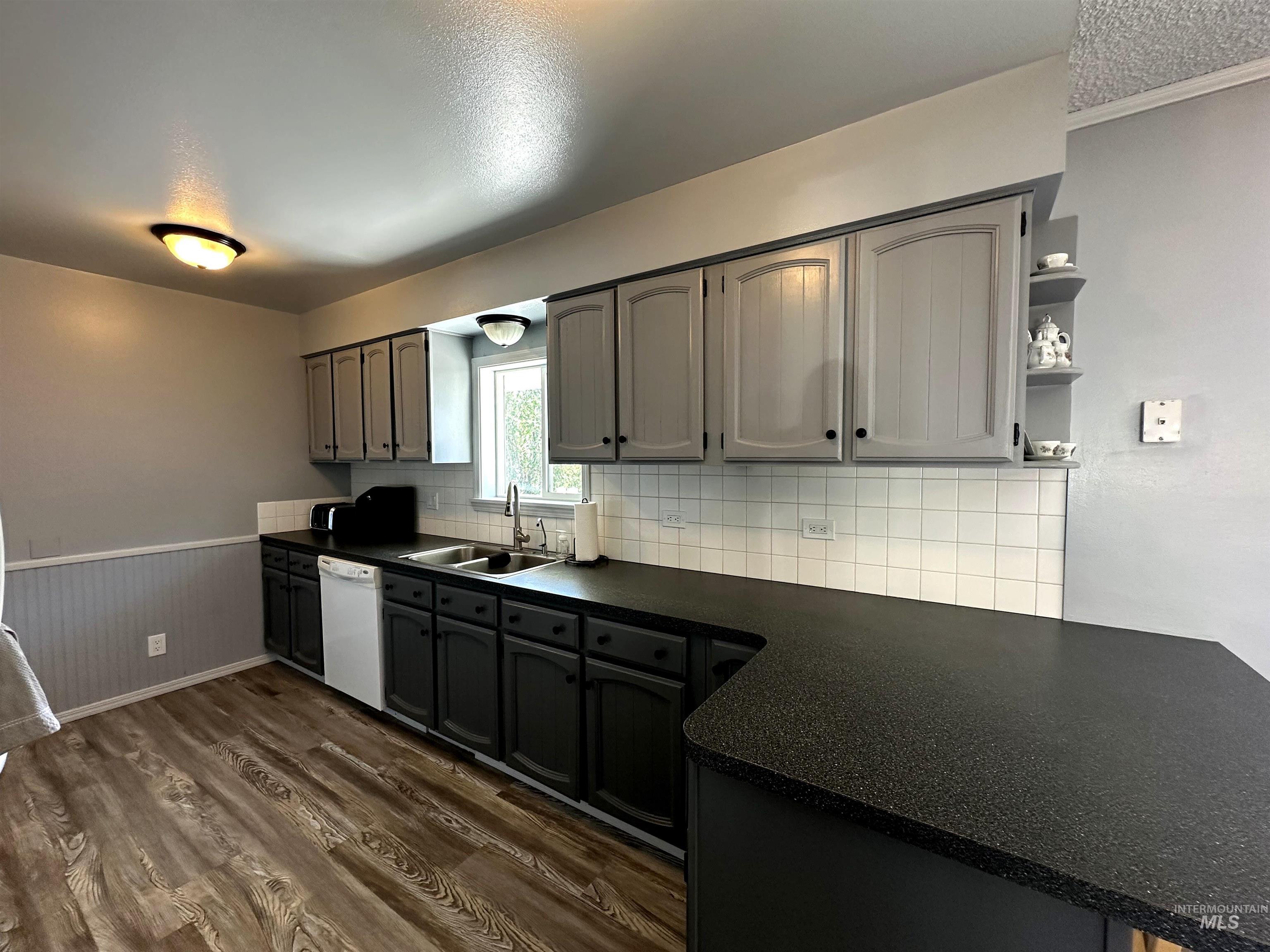 Kitchen featuring white dishwasher, dark countertops, dark wood-type flooring, a wainscoted wall, and tasteful backsplash