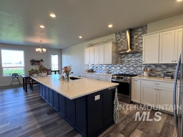 Kitchen featuring backsplash, stainless steel gas range, a kitchen island with sink, wall chimney exhaust hood, and white cabinetry