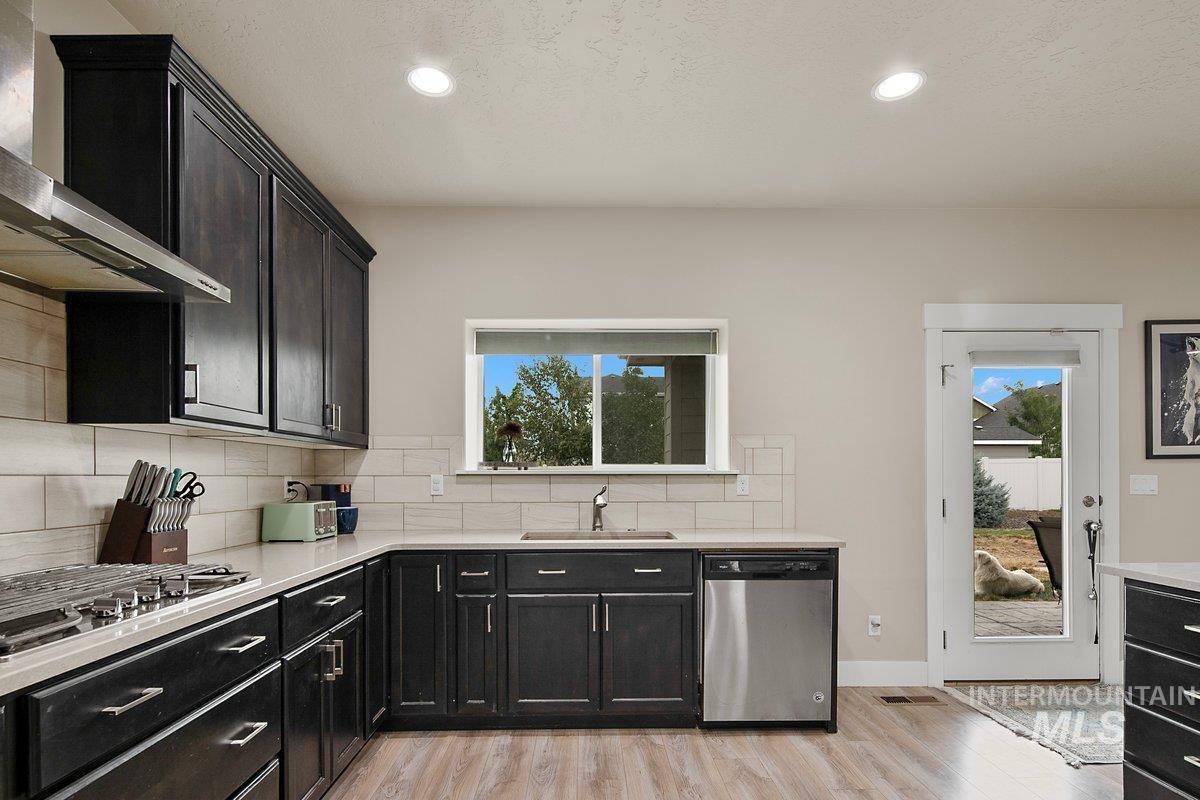 Kitchen with dark cabinets, wall chimney range hood, light wood-style flooring, appliances with stainless steel finishes, and backsplash