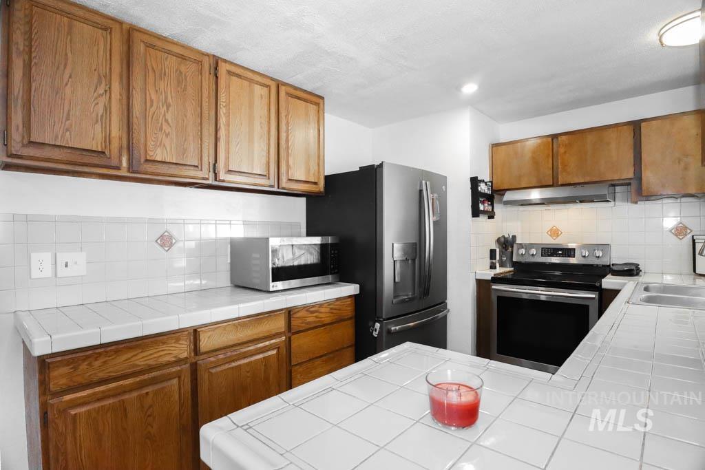 Kitchen featuring appliances with stainless steel finishes, backsplash, brown cabinets, under cabinet range hood, and tile countertops