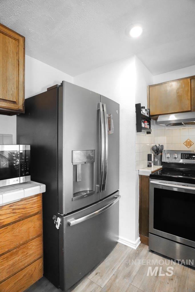 Kitchen with brown cabinets, appliances with stainless steel finishes, decorative backsplash, light wood-style floors, and a textured ceiling
