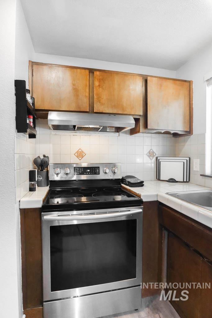 Kitchen featuring tile countertops, stainless steel range with electric stovetop, decorative backsplash, under cabinet range hood, and brown cabinetry