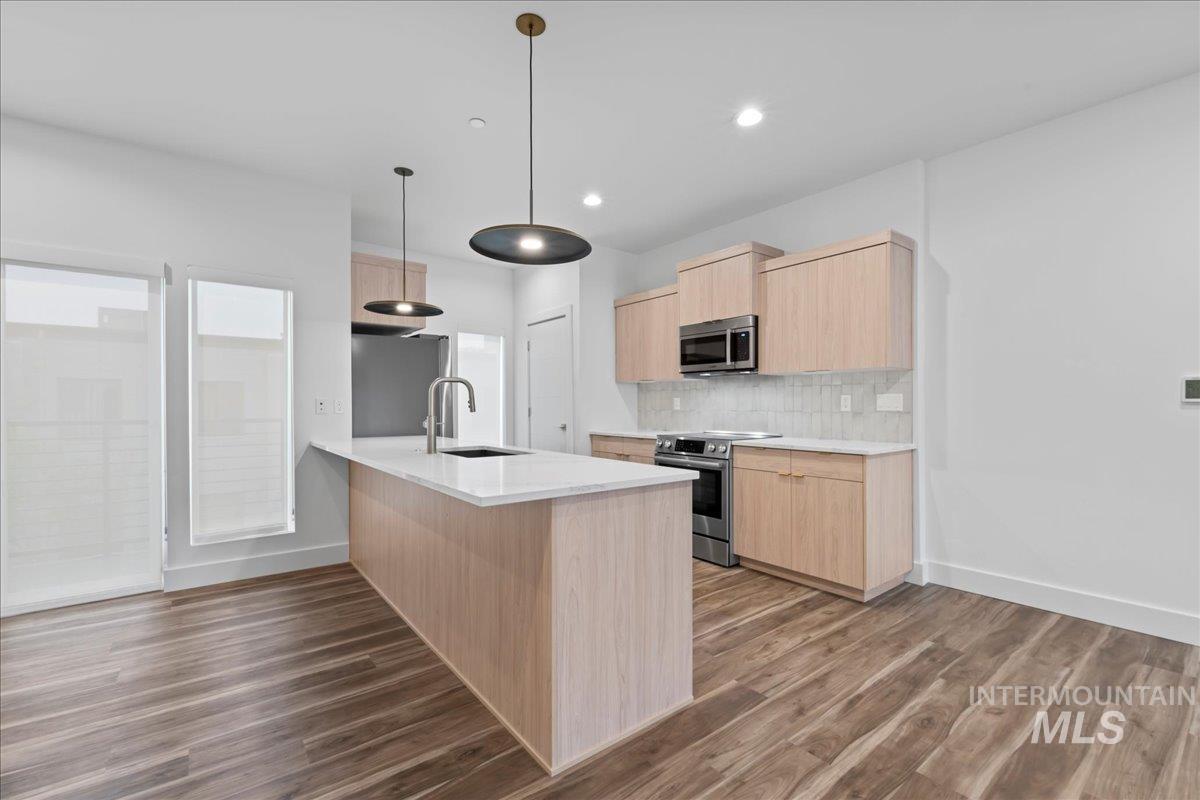 Kitchen featuring light brown cabinetry, stainless steel appliances, backsplash, light stone counters, and hanging light fixtures